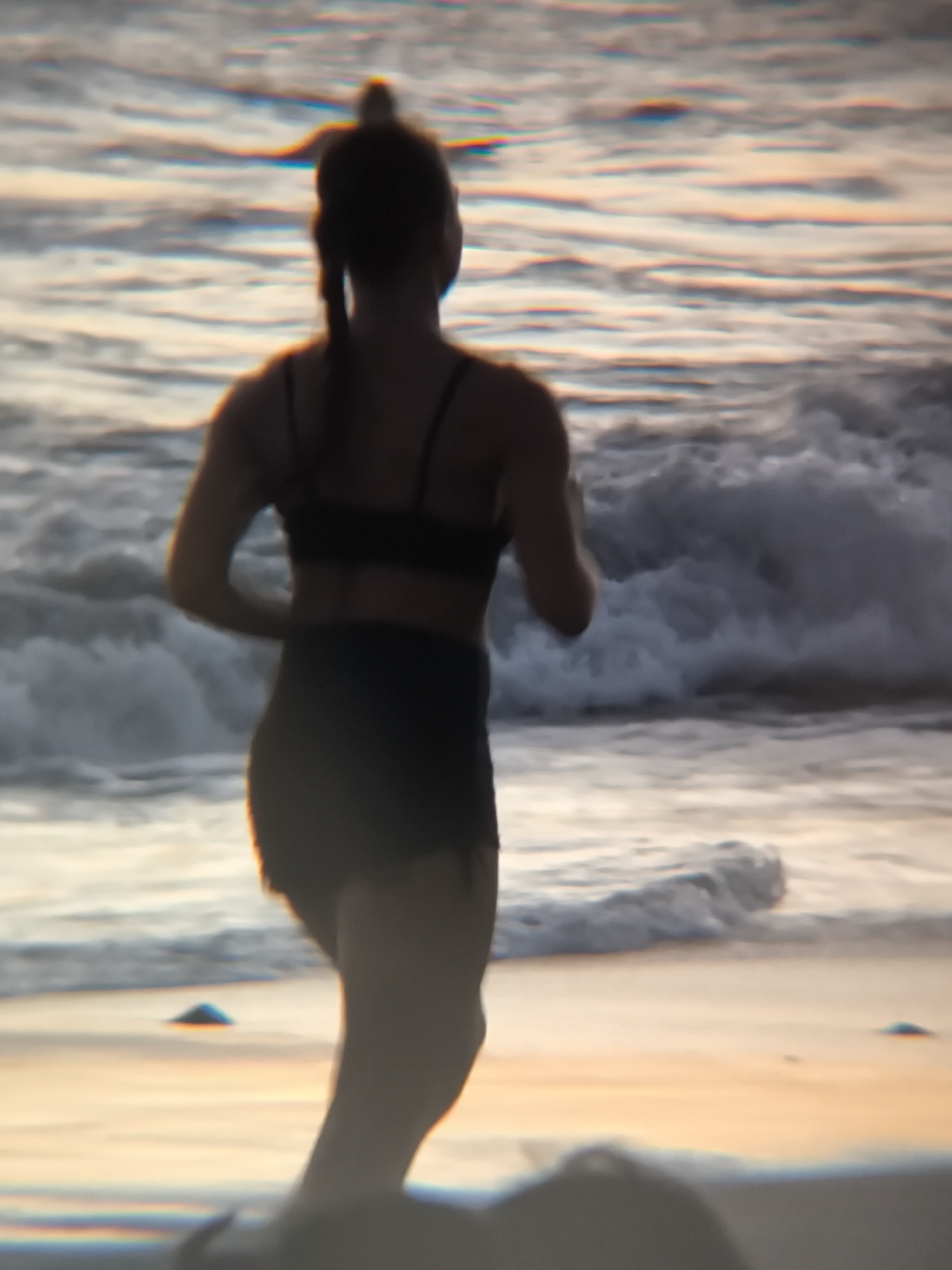 A woman with her hair in a ponytail is running along the beach, facing away from the camera. She is wearing a black sports bra and black shorts. The beach setting includes sand and breaking waves. The location is the city of North Kuta, Indonesia.
