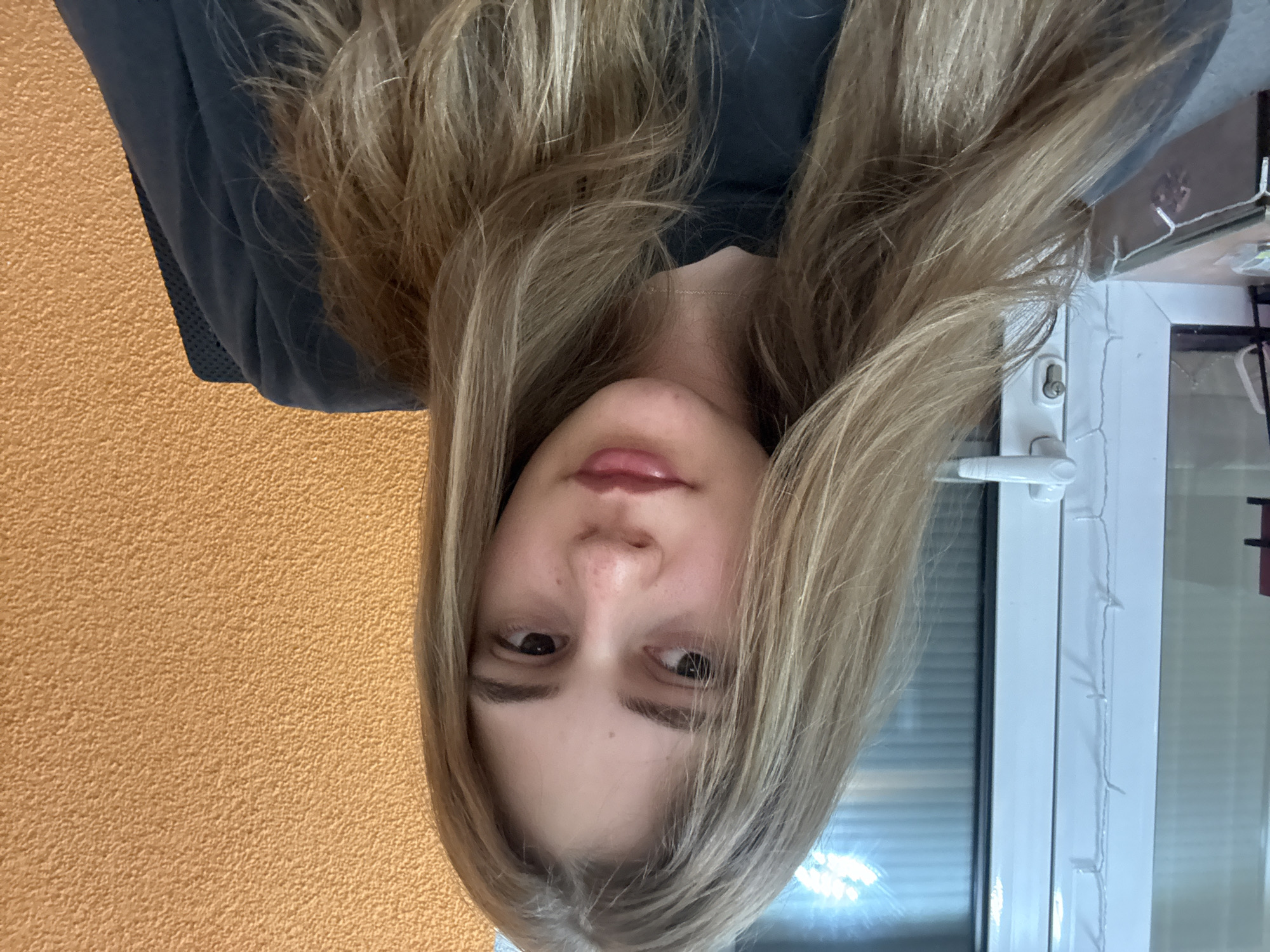 A young woman with light blonde hair is centered in the image, facing the camera. The background consists of a textured orange wall on the left and a white window frame and window with blinds on the right. The woman wears a dark grey top. A thin gold necklace is visible around her neck. The image is rotated 180 degrees. There is no indication of this image being taken in Panagyurishte, Bulgaria.