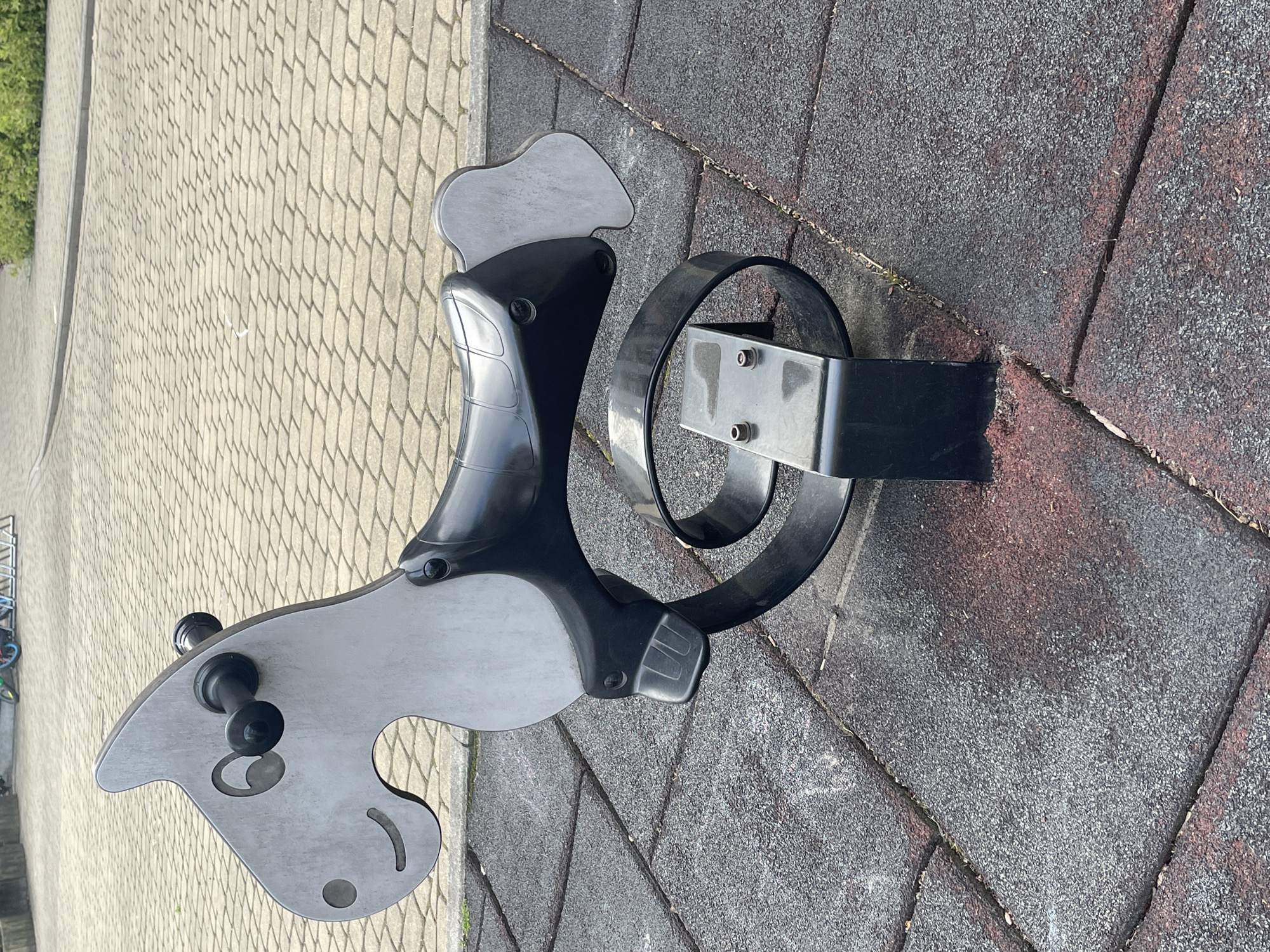 The image depicts a child's playground spring rider shaped like a horse. The horse figure is constructed from gray and black metal, with a saddle and reins. It is bolted to a black metal spring base that is attached to the ground. The playground surface surrounding the spring rider consists of interlocking gray and reddish-brown tiles. To the left, a section of cobblestone pavement is visible, with greenery in the background, suggesting a park or public space.