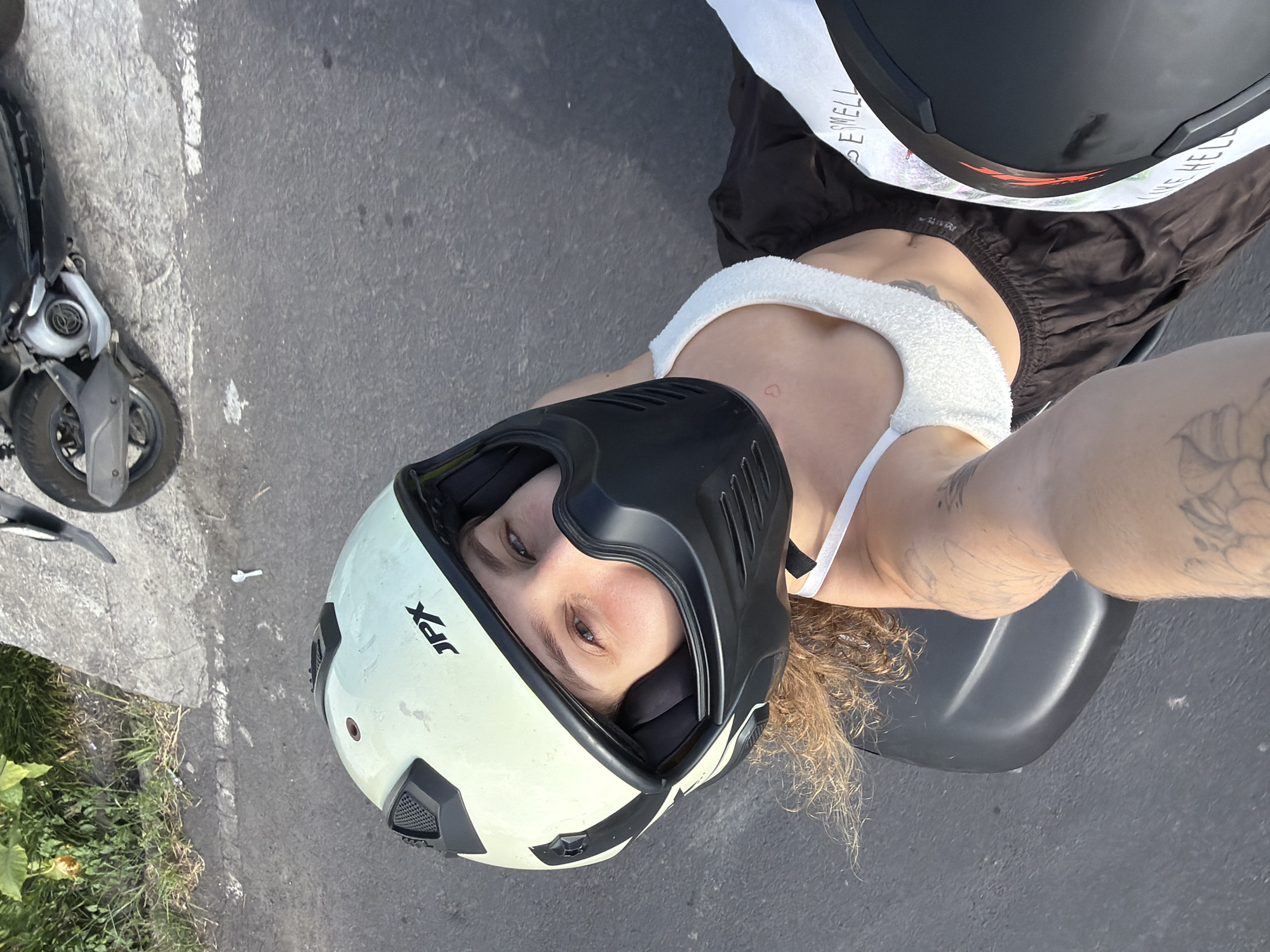 This media file shows a young woman wearing a white and black helmet, lying on the ground next to a scooter. The scooter is parked on an asphalt surface, with some grass and foliage visible to the left. The woman is wearing a white tank top and dark pants. She has tattoos on her arm. The helmet is a full-face helmet with a visor. Visible text on the helmet includes "JPX" and some unreadable lettering on a white strap. The scene appears to be outdoors during the daytime, with sunlight casting shadows. The woman's expression is difficult to ascertain due to the helmet, but her eyes are visible and seem to be looking upwards. The overall impression is that she is taking a break or posing for a photo after riding her scooter. The location context of North Kuta, Indonesia suggests a warm, tropical environment, which aligns with the outdoor setting and the woman's attire.
