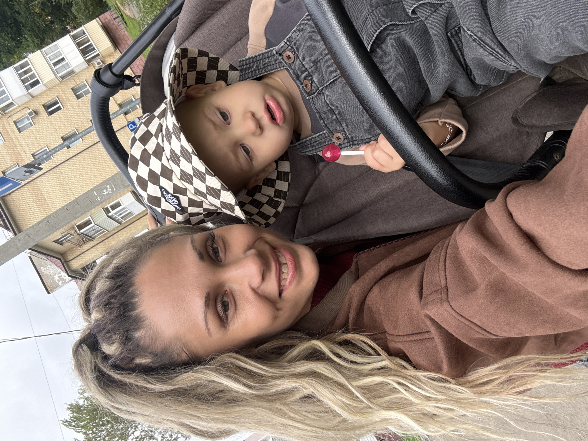 A medium shot selfie shows a woman and a young child in a stroller, set against a backdrop of a city street in Rusanovo, Russia. The woman, with blonde hair styled in a high ponytail, smiles at the camera, her face framed by a brown jacket. Beside her, the child, wearing a checkered hat and denim overalls, sits in the stroller holding a red lollipop. In the background, apartment buildings and trees line the street under an overcast sky. The scene appears to be captured during a casual outing.