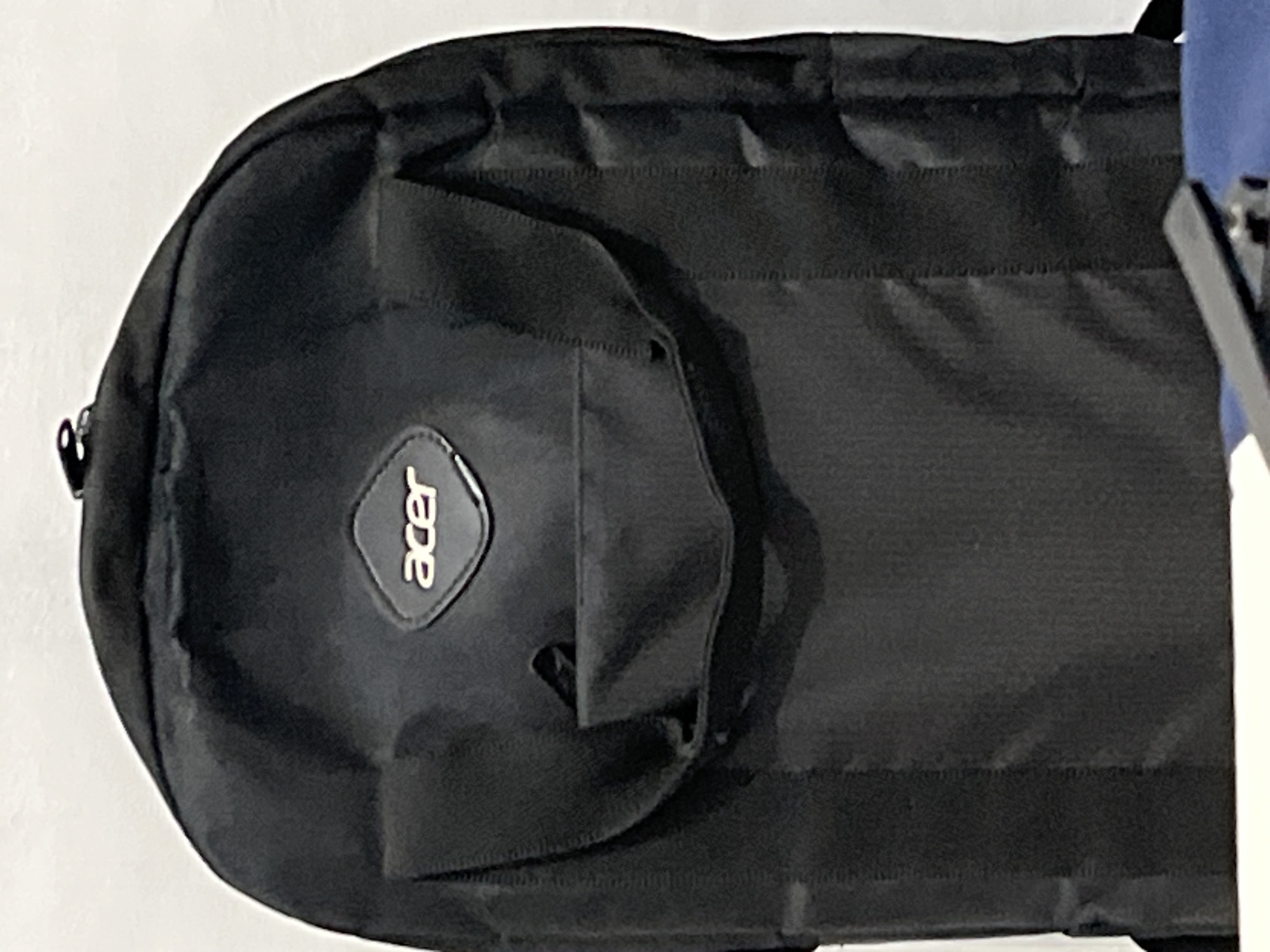 A black soft-sided bag, likely a laptop case or a small travel bag, is depicted in a vertical orientation. The bag is made of a matte, possibly synthetic, fabric with visible stitching and a structured, somewhat padded appearance. A distinctive, silver-colored "acer" logo, rendered in its signature lowercase stylized font, is prominently featured on a black, slightly raised, tear-drop shaped patch on the front of the bag, towards the upper left when viewed upright. Below the logo, a flap or small pocket is visible. The bag features horizontal stitching lines, suggesting multiple compartments or design elements on its front surface.
The background is a plain, light-colored surface, possibly a wall or table. To the right edge of the frame, a sliver of a blue object and a white vertical edge are visible, though their nature is indistinct. No specific activity is taking place; the bag appears to be stationary. The lighting suggests an indoor setting, but the time of day, weather, or any emotional context cannot be determined from the image. The image itself contains no visual cues to confirm its location in Lhokseumawe, Indonesia, beyond the provided context.