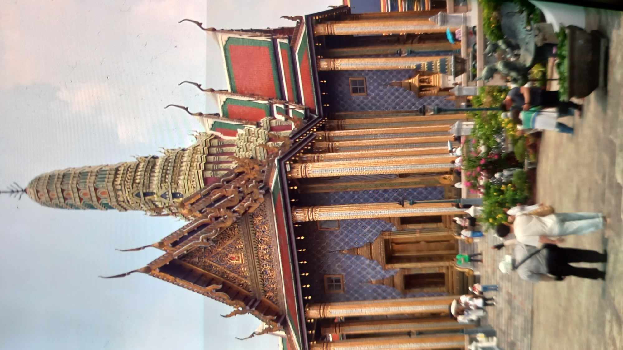 The image shows a view of a temple with intricately decorated spires and roofs, featuring a predominant color palette of gold, red, green, and blue. Several people are visible in the scene, some walking and others standing, appearing to be tourists or visitors. The setting is an outdoor paved area in front of the temple. Lush green plants and colorful flowers are interspersed throughout the foreground, adding a vibrant touch to the scene. The lighting suggests it is daytime, possibly with some cloud cover. The overall atmosphere seems to be one of casual observation and exploration of the ornate architecture and gardens. There is no visible text in the image. The location context provided is ตำบลเกาะพะงัน, Thailand, which is a district on the island of Koh Phangan in Surat Thani province.