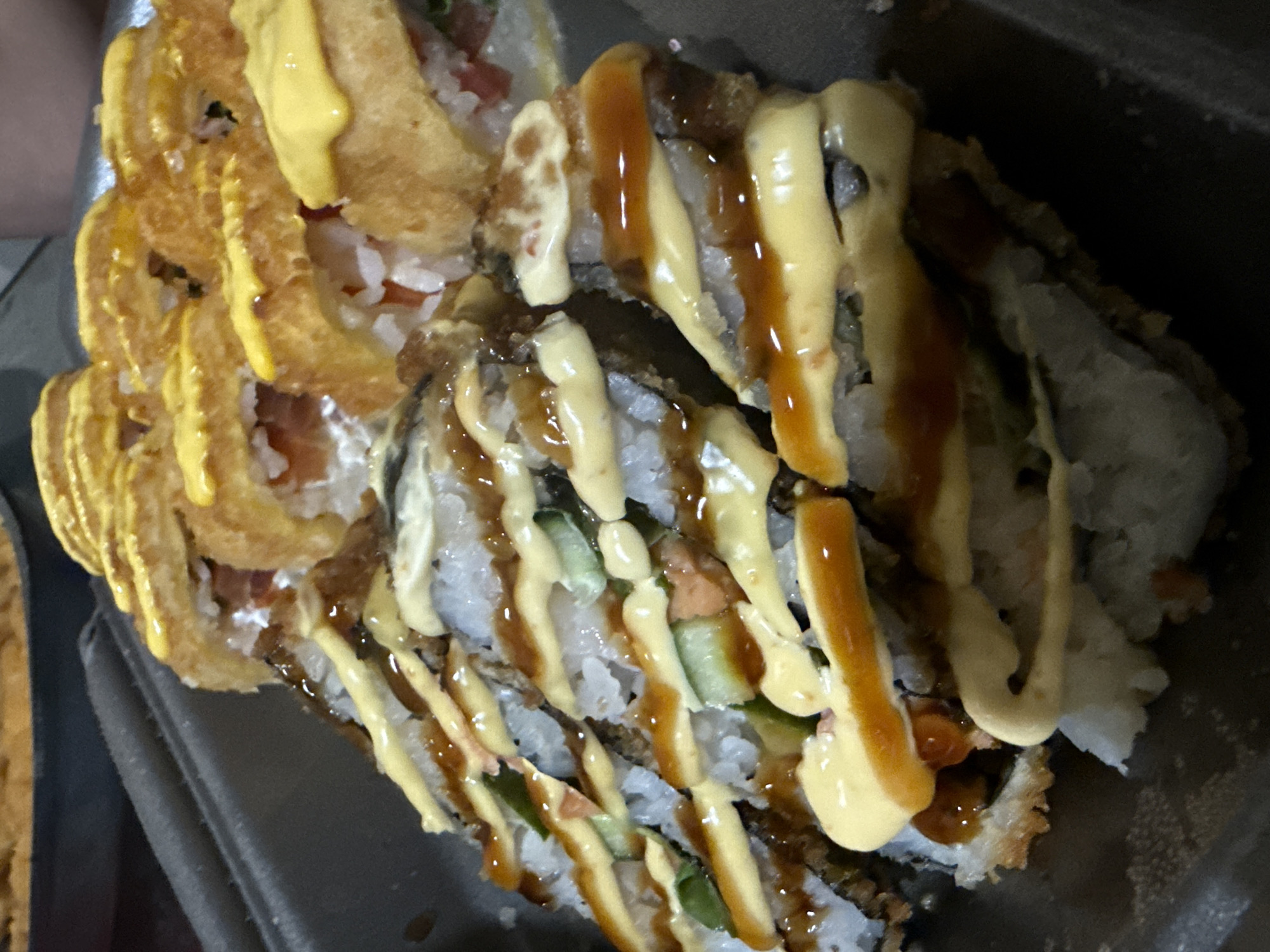 The image shows a close-up of sushi rolls in a dark container. The sushi appears to be topped with sauces, one a light yellow and the other a dark brown. Some rolls have a crispy topping. The image provides no information about the city of Dmitrov, Russia, or any people, actions, or interactions. The environment is limited to the interior of the container holding the sushi.