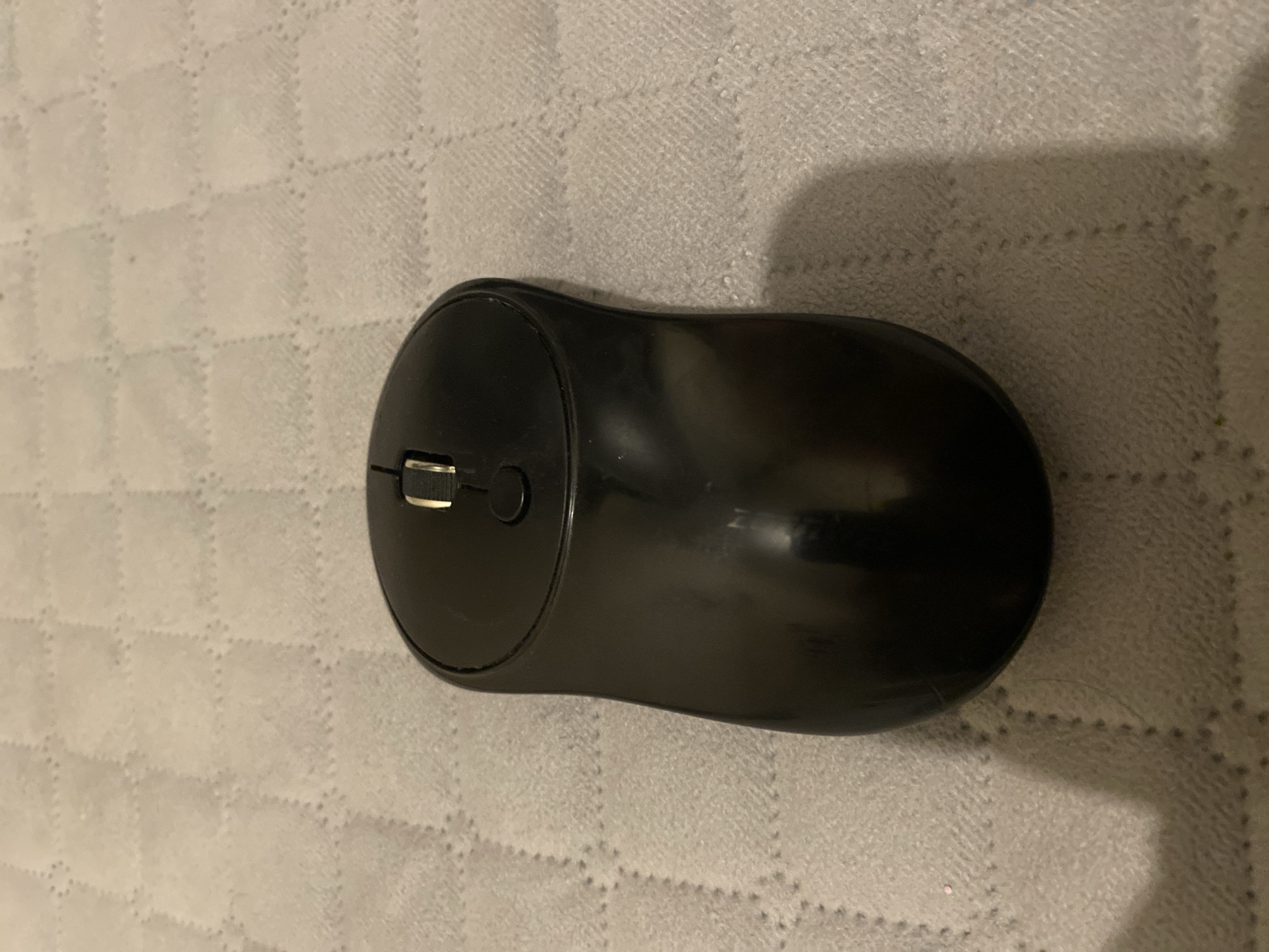 A black wireless computer mouse is centered on a textured light gray surface, possibly a blanket or cloth. The mouse is oriented vertically with its scroll wheel at the top. The surface beneath the mouse has a quilted pattern of small, diamond-shaped indentations. There are no people or discernible activity in the image. The lighting appears to be from an overhead source, casting a slight shadow to the right of the mouse. The scene is indoors and does not contain any information to suggest a specific location in Kizilyurt, Russia.