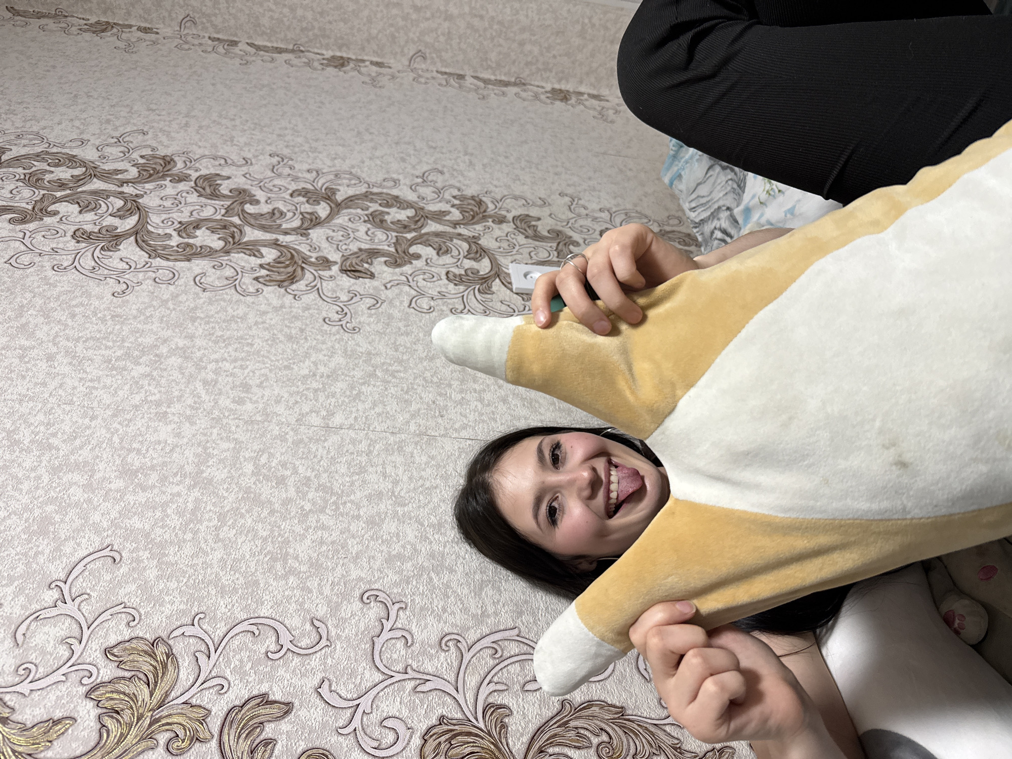 A woman with dark hair lies on a bed with a patterned beige bedspread. She is holding a large plush pillow shaped like a corgi dog over her face and smiling with her tongue sticking out. Part of a person wearing black pants is visible in the upper right corner. A white electrical outlet is visible on the bedspread near the woman's hand. The image appears to be taken indoors. The city of Фоки is not discernible from the image.