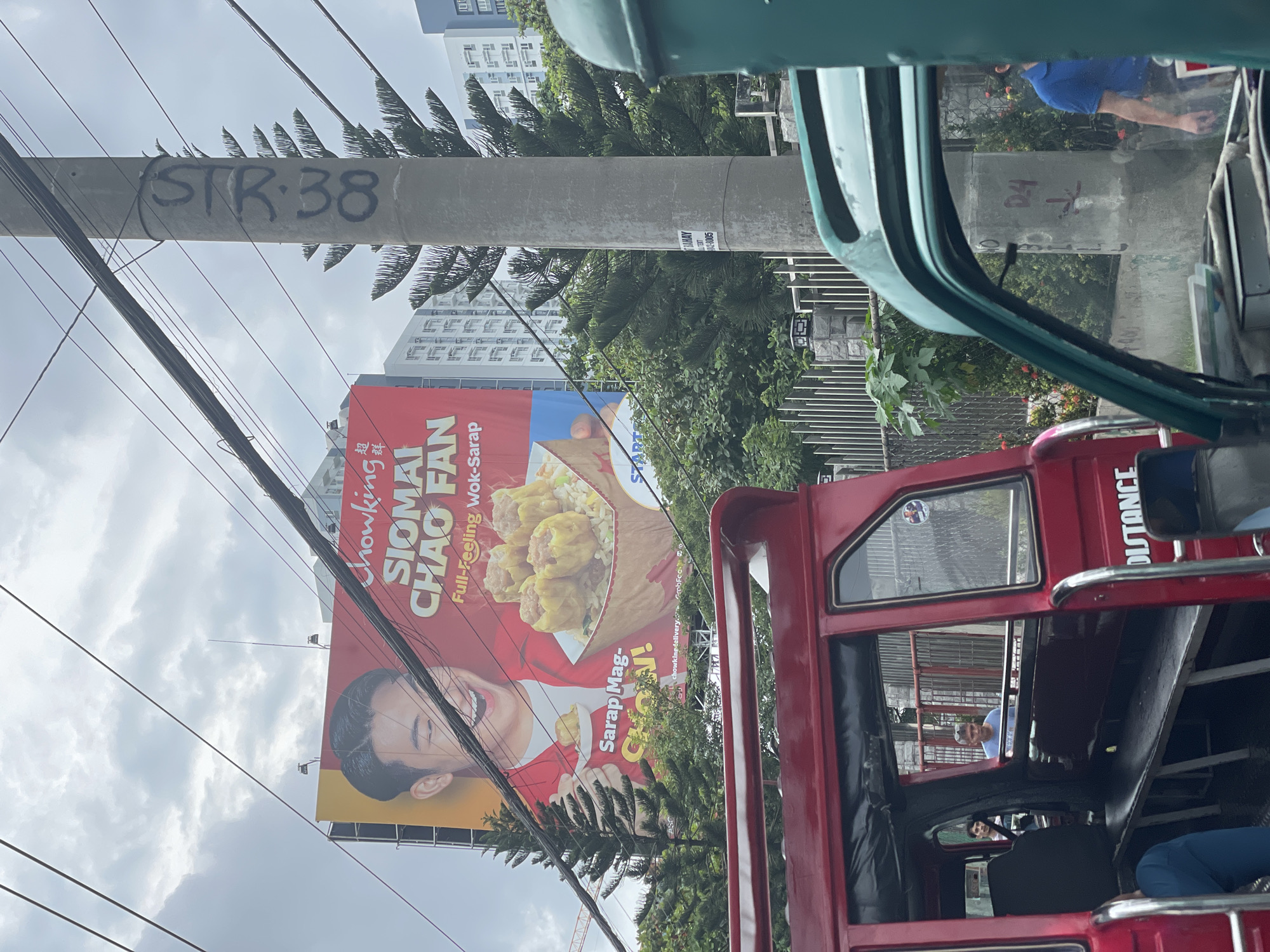 A billboard advertising Chowking's Siomai Chao Fan is visible in the upper portion of the image. The billboard features a smiling man, a serving of siomai and fried rice, and text in English and Chinese. Below the billboard, a concrete utility pole with graffiti reading "SR 38" is present. In the foreground, a red, open-air vehicle resembling a jeepney or tricycle is parked, with people visible inside and near it. A green vehicle is partially visible on the right side of the frame. The scene appears to be on a street in Davao City, Philippines, indicated by the context of Chowking, a popular fast-food chain in the Philippines, and the presence of local vehicles and urban infrastructure. The sky is overcast.