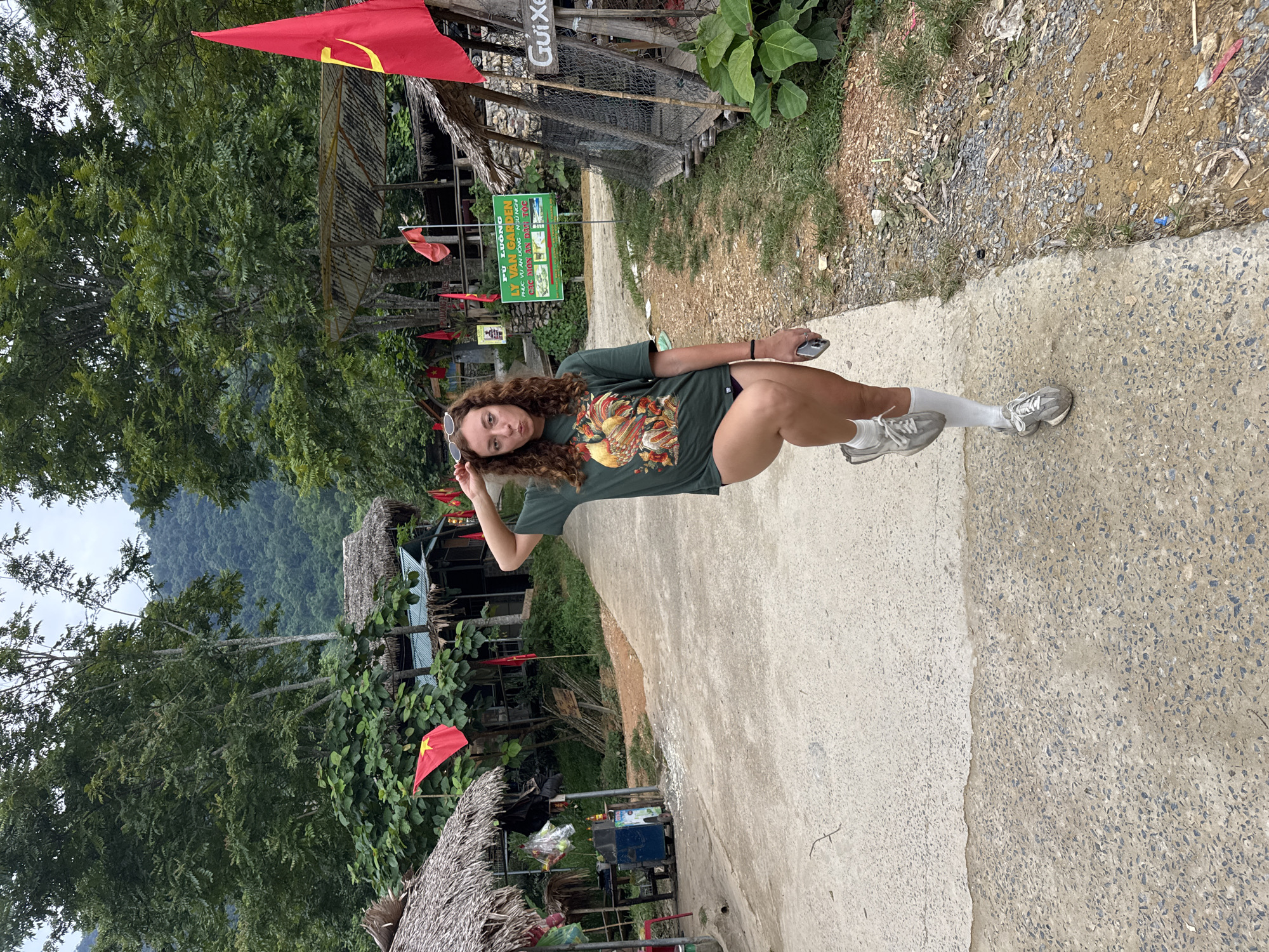 A young woman with curly brown hair is standing on a concrete path in what appears to be a rural Vietnamese village. She is wearing a dark green t-shirt with a colorful graphic of a rooster and some floral elements, dark shorts, white socks, and grey athletic shoes. She is holding a silver smartphone in her right hand and has her left hand resting on her head.
The setting is lush and green, with trees and vegetation visible in the background, along with some simple structures that appear to be thatched-roof huts or stalls. Red flags with yellow stars, likely Vietnamese flags, are present, some attached to poles and others flying from buildings. A sign with Vietnamese text, including "PÙ LUÔNG" and "LY VAN GARDEN," is visible, suggesting a tourist or hospitality area. The path is made of concrete and has some gravel and dirt alongside it. The overall impression is of a warm, possibly humid day in a scenic, natural environment.