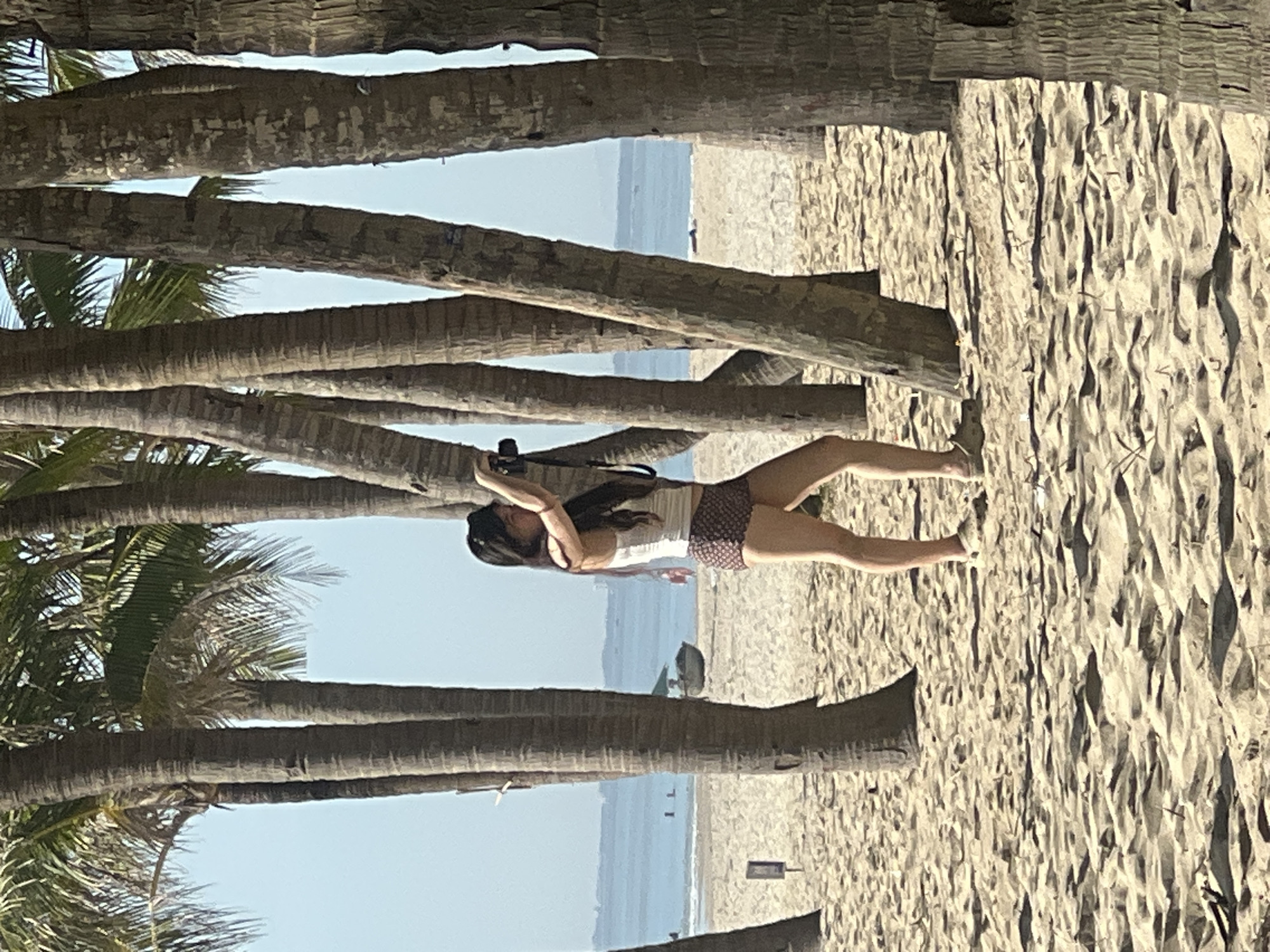 The image shows a woman standing on a sandy beach, surrounded by palm trees, holding a camera. She is wearing a white tank top and patterned shorts. The background reveals the ocean and a clear sky, with some distant boats. The location is likely a beach in Da Nang, Vietnam, known for its palm-lined coastlines. The scene captures a moment of leisure and photography amidst a tropical environment.