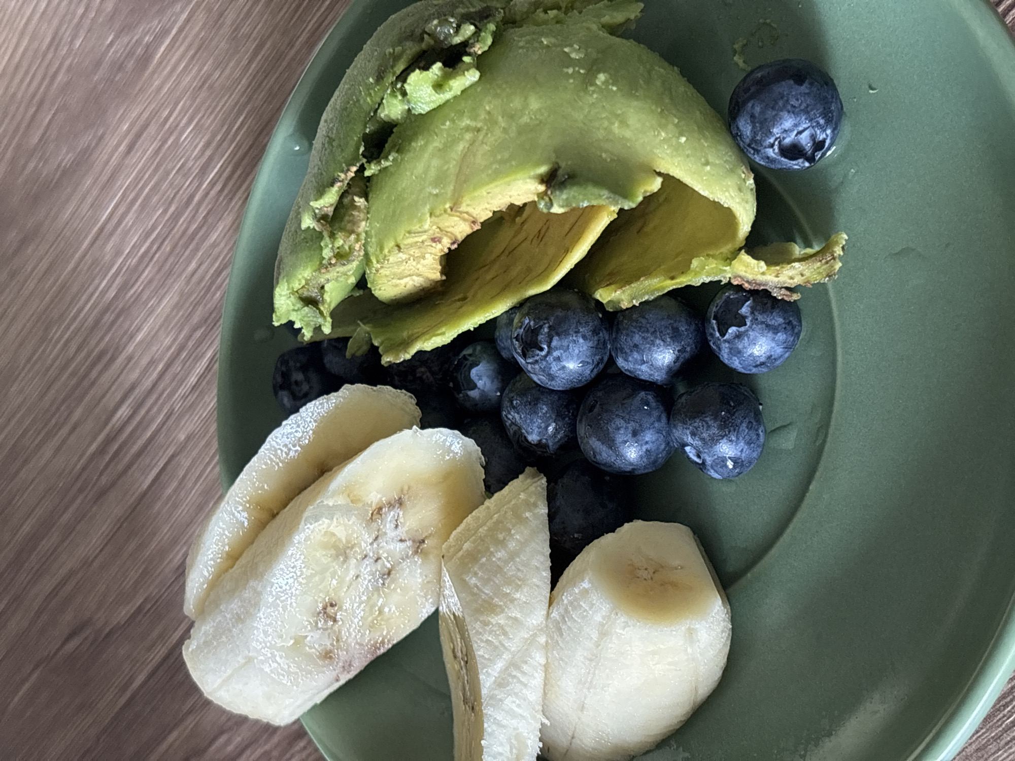 A close-up shot shows a green plate on a wooden surface. The plate contains a peeled and slightly distressed avocado with some flesh still attached to the skin, a cluster of blueberries, and slices of banana. The scene appears to be indoors, possibly in a dining or kitchen area. There is no indication of the location being in Choeng Thale, Thailand, or any people or actions visible in the frame.