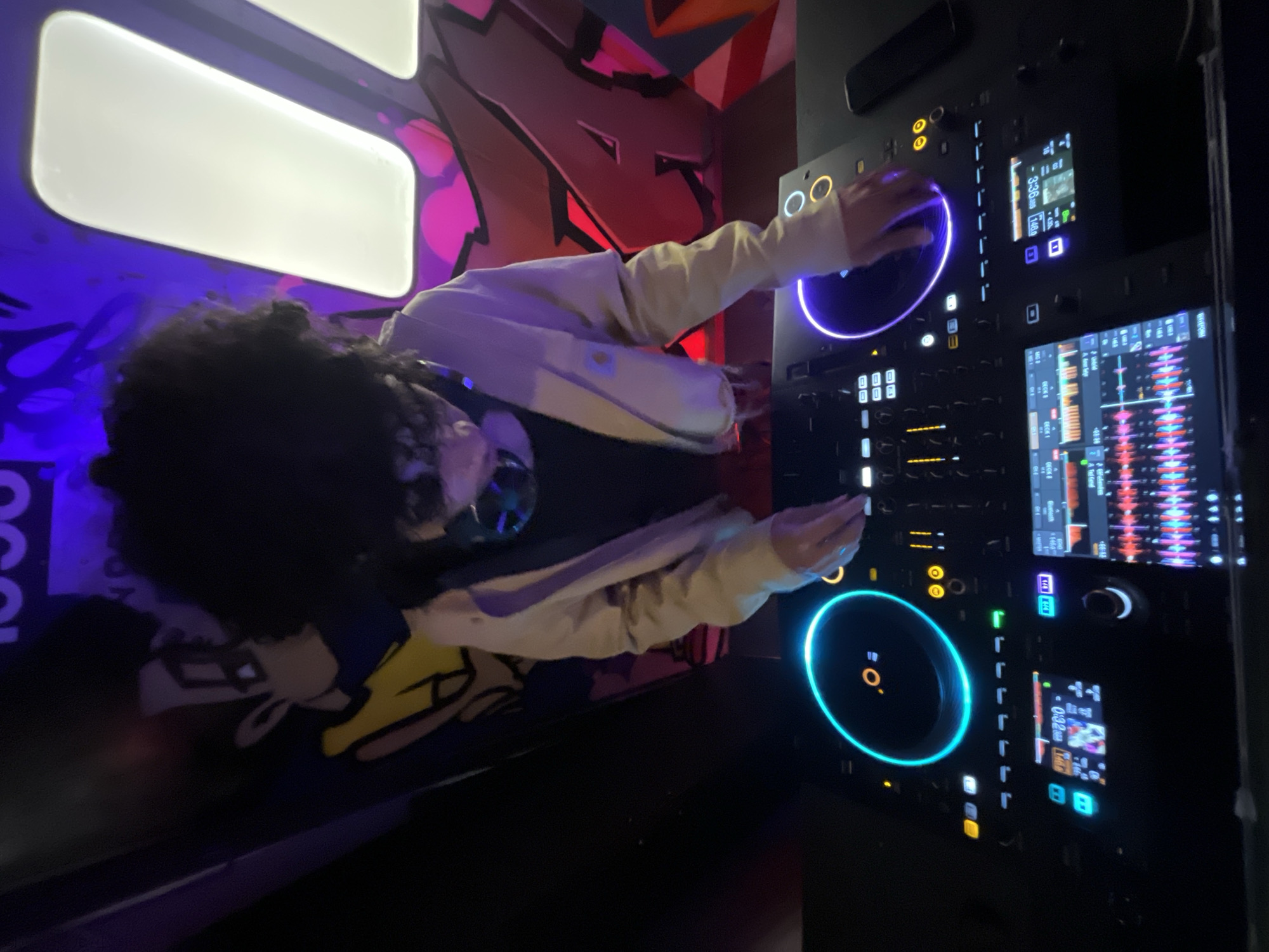 A dimly lit club scene shows a DJ working at a professional DJ deck. The DJ, wearing a light-colored jacket, stands behind the equipment with hands positioned over the turntables, suggesting an active performance. The DJ has dark curly hair and wears sunglasses. The deck itself is black with illuminated buttons, screens displaying waveforms, and bright neon-blue-lit jog wheels. Graffiti art adorns the walls, featuring vibrant colors and abstract shapes, adding to the club's aesthetic. The venue appears to be in Barcelona, Spain. The photograph is taken at an angle, presenting a slightly tilted view of the scene.