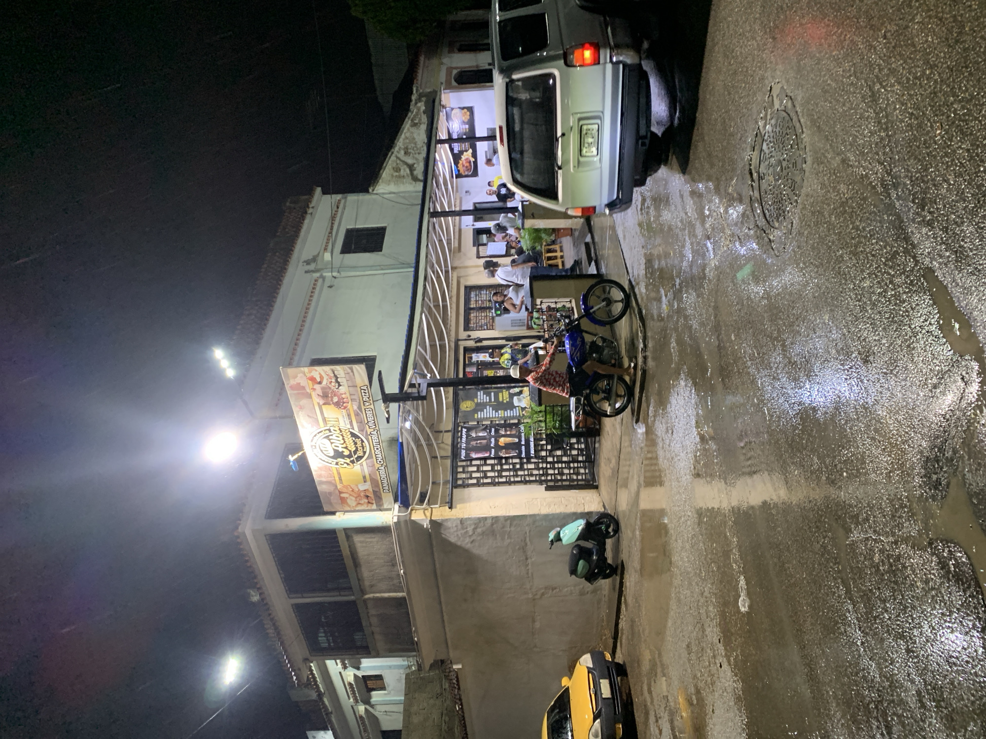 The image depicts a nighttime scene on a wet street in Santa Ana, Venezuela, likely in front of a food establishment. Rain is visible falling, contributing to the reflections on the asphalt. Several vehicles are present: a yellow car parked on the left, a light blue scooter, and a silver SUV facing away from the viewer on the right. A person is riding a blue motorcycle towards the viewer.
The building in the background appears to be a business with a prominent sign reading "El Alba Market" which also advertises "Panadería, Charcutería, Víveres y Pizza." Inside, under an awning, several people are gathered, some standing and some seated at tables. Men and women are visible, engaged in conversation or ordering food. The establishment has large windows and an open-air seating area. The street is illuminated by artificial lights from the business and streetlights, creating a bright, slightly hazy atmosphere. A manhole cover is visible in the wet street.