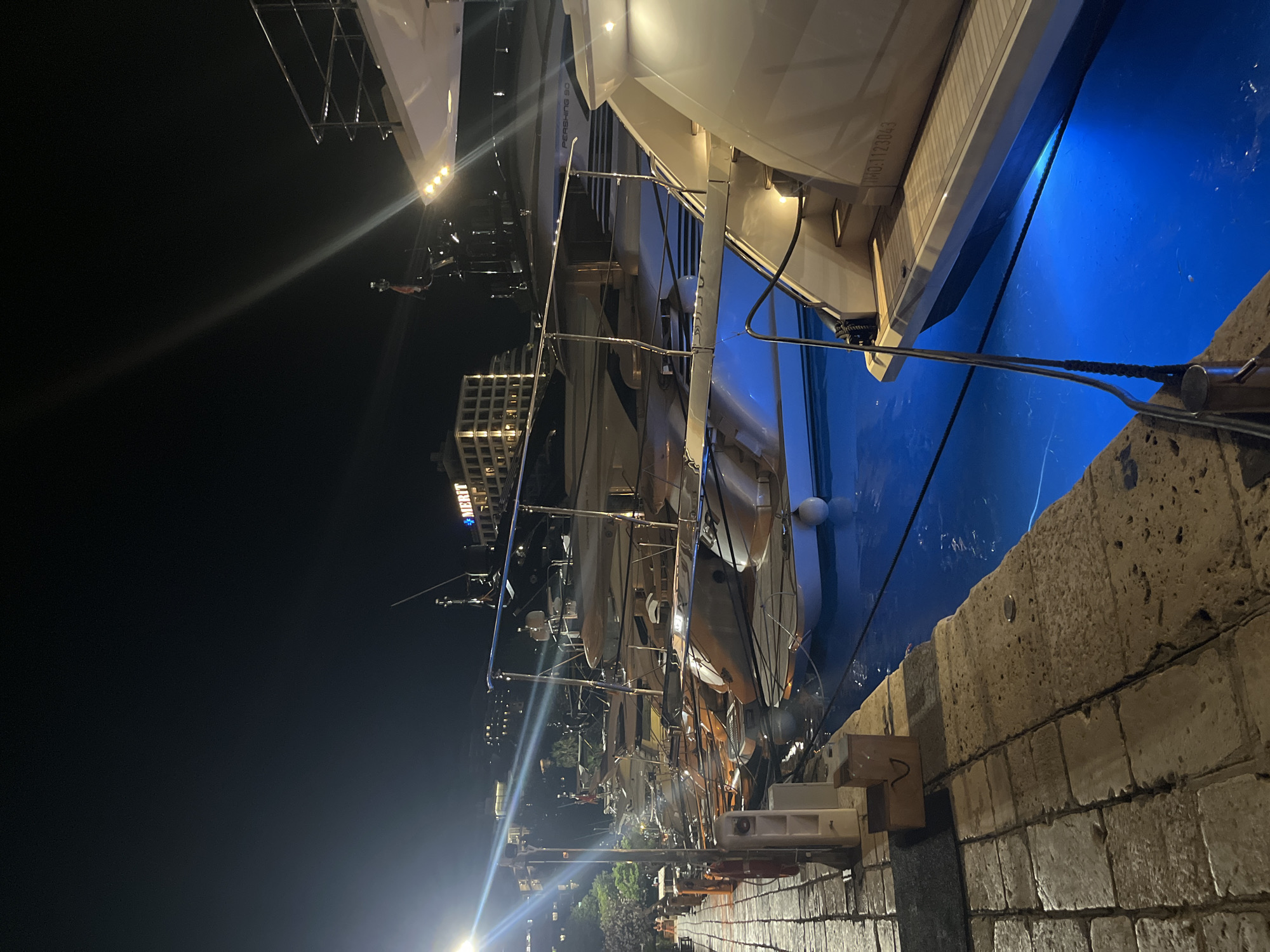 The image depicts a night scene at a marina in Budva, Montenegro, likely the Budva Marina. Several yachts are docked along a cobblestone promenade. The water beneath the yachts is illuminated with bright blue lights, creating a vibrant contrast with the dark night sky. In the background, a building with illuminated windows is visible, featuring a sign that reads "MERIT". The promenade is lined with bollards and ropes used for securing the vessels. A few individuals can be seen on the far left of the promenade, suggesting a low level of activity. The overall atmosphere is tranquil and luxurious, characteristic of a marina at night.