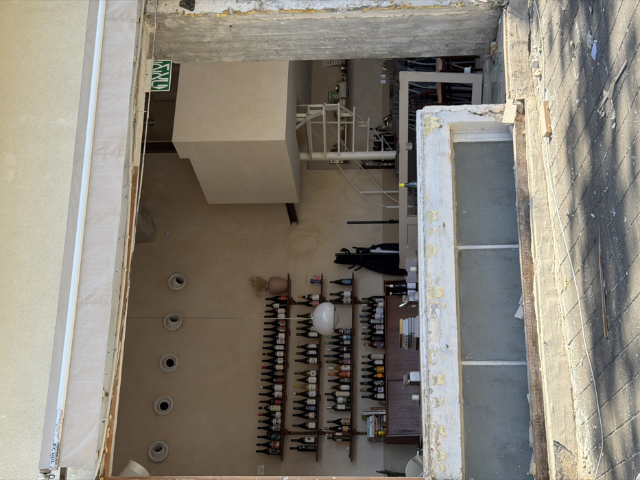 An exterior view captures the open entrance of a commercial establishment, likely a bar or wine shop, in Tel-Aviv, Israel. The ground in the immediate foreground is paved with dark grey rectangular tiles, strewn with white debris, thin wires, and a piece of cardboard. A heavily damaged and peeling off-white lower facade, possibly a window frame, occupies the center foreground. To the far right, a rough concrete pillar with exposed aggregate borders the scene.
Above the entrance, a light-colored retractable awning displays Hebrew text ("הכל על הבר").
Inside the establishment, a light beige-colored wall extends across the back. Mounted on this wall are four wooden shelves, each displaying numerous dark-colored bottles, predominantly wine or spirits. A large white spherical pendant light hangs in front of the middle shelves. To the right of the shelves, a small pink vase holds dried flora. Below the shelves, a dark wooden counter or bar is visible, topped with stacks of books, additional bottles, and glasses. A black coat rack stands to the right of the counter.
Towards the rear right of the interior, a large, light-colored rectangular structure, possibly a mezzanine or architectural feature, extends from the ceiling. Beneath it, a white metal spiral staircase descends. Below the staircase, a seating area with tables and chairs, some with red upholstery, is partially visible. A green exit sign with white Hebrew text ("יציאה") is mounted high on the right wall. The interior appears unoccupied, and the state of the entrance suggests ongoing renovation or prior damage.