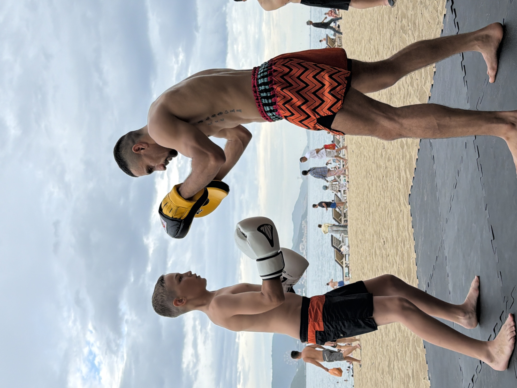 Two males, one adult and one juvenile, are engaged in a boxing or kickboxing training session on a sandy beach in Nha Trang, Vietnam. The adult, positioned to the right, wears orange and black chevron-patterned shorts and yellow training mitts on his left hand. He has a muscular physique and a tattoo on his torso. The juvenile, to the left, wears black shorts with an orange waistband and white boxing gloves. He is looking towards the adult. They are standing on a black mat on the sand. In the background, the ocean is visible with calm water and a cloudy sky. Several beachgoers and lounge chairs are scattered along the shoreline, with a cityscape and hills in the distance. The overall atmosphere suggests an outdoor training activity in a coastal urban environment.