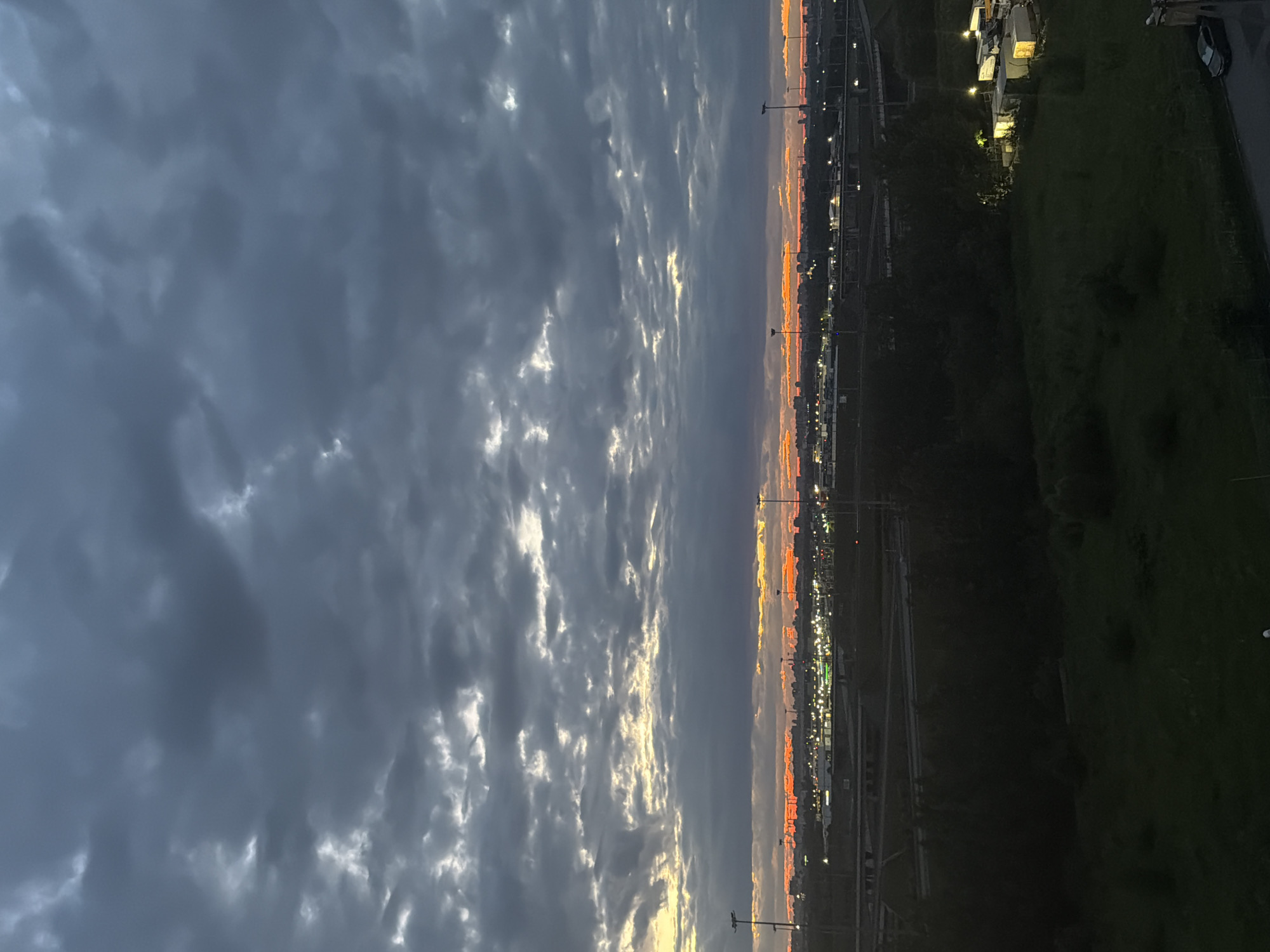 An image captures a dusky sky over a city, likely at dusk or dawn. The sky is filled with heavy, dark gray clouds, interspersed with patches of lighter, illuminated clouds that suggest the presence of the sun below the horizon. A band of vibrant orange and yellow hues stretches across the horizon, indicating the sun's position.
Below the sky lies a cityscape dominated by a dark silhouette. In the mid-ground, an industrial or commercial area is visible with numerous lights illuminating buildings and infrastructure, including what appear to be streetlights or floodlights. Roads or highways with overpasses are also discernible in this area. Further in the foreground, a dark, overgrown area of vegetation, possibly a field or park, transitions into a dark expanse, implying the presence of trees or a densely vegetated area. A few scattered lights from structures or vehicles are visible within this dark foreground. In the lower right corner, a dark-colored car is parked on a paved surface, adjacent to what might be a building or structure. The overall scene is characterized by low light conditions and a somewhat ominous or serene atmosphere due to the dramatic sky and the dim lighting of the city.