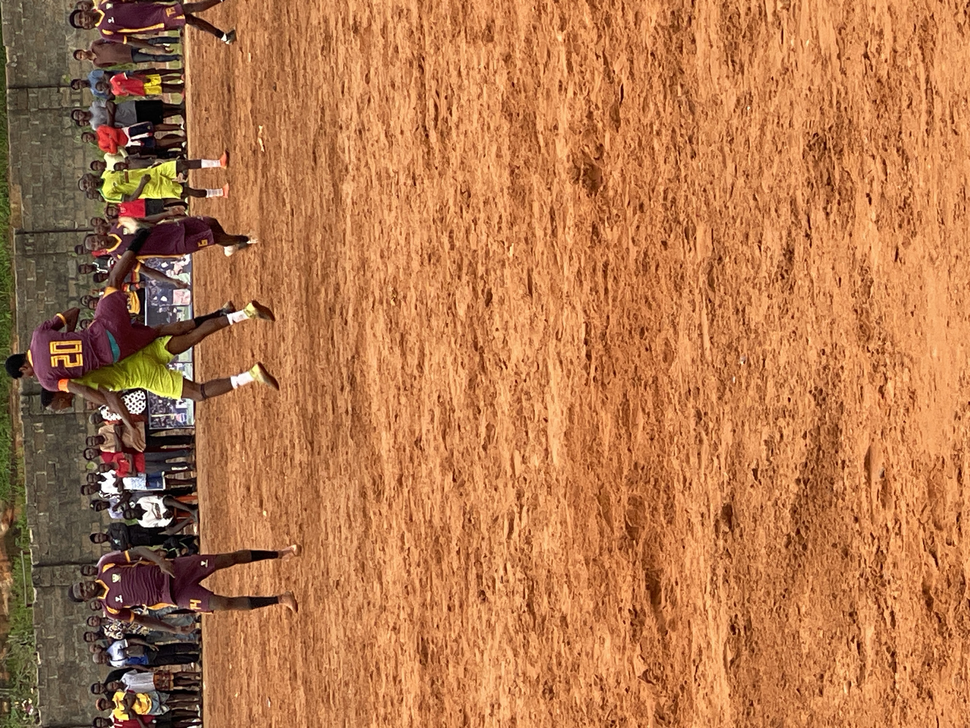 The image depicts a soccer match being played on a dirt field in an outdoor setting. In the foreground, a player in a maroon and gold uniform is in mid-air, attempting to head a soccer ball. He is wearing a number 4 on his shorts. Another player in a lime green uniform is also in the air, seemingly attempting to block the header. He is wearing bright orange socks. Behind these two players, other participants and spectators are visible. Spectators are standing and sitting along the edge of the field, with a brick wall behind them. The ground is a reddish-brown dirt, uneven and textured. The overall scene suggests a casual or local soccer game.