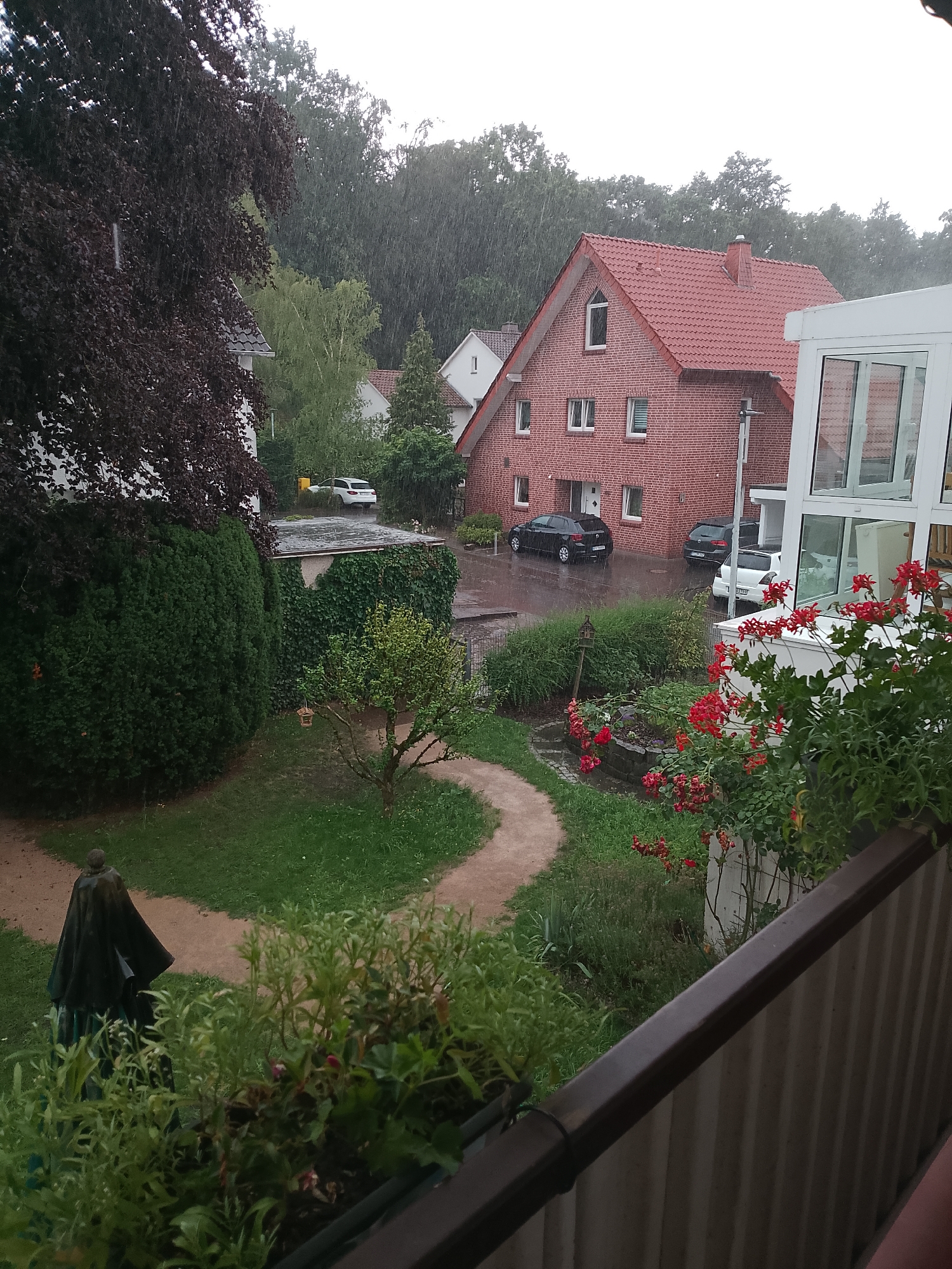The image depicts a residential street in Rheda-Wiedenbrück, Germany, during a heavy rain shower. The foreground is dominated by a railing and lush green plants, including flowering red geraniums, suggesting a balcony or elevated viewpoint. Beyond this, a well-maintained garden features a winding path, a small tree with a bird feeder, and a dark green statue of a cloaked figure.
Further into the scene, several cars are parked on a wet driveway and street, including a white SUV, a black sedan, and a silver car. Two brick houses with red tiled roofs are visible, one larger with a prominent chimney, and another smaller white house. The surrounding area is densely populated with trees and hedges, all appearing damp from the rain. The sky is overcast, contributing to the gloomy, wet atmosphere. No people are clearly visible in the image.