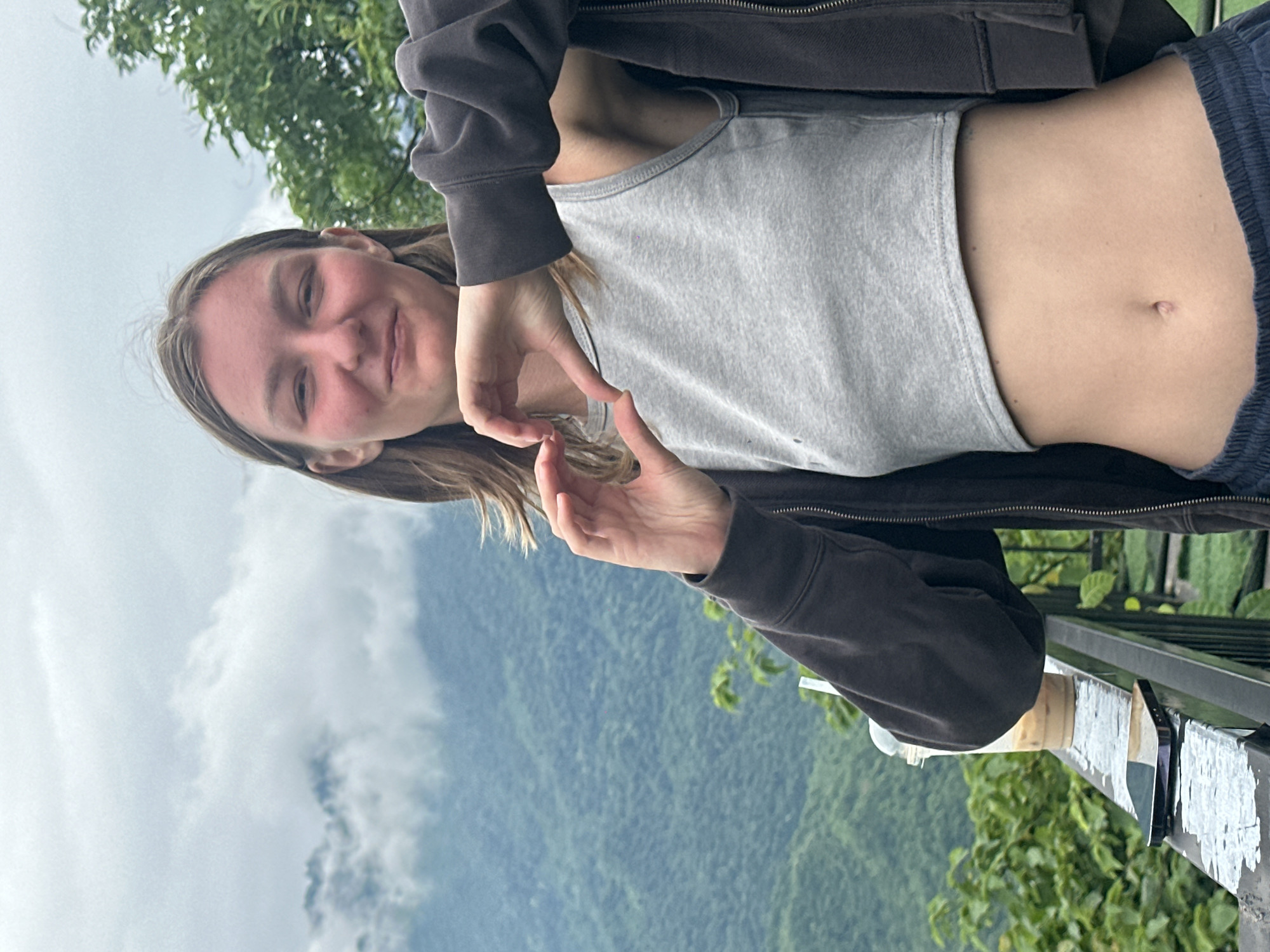 A young woman with long, light brown hair smiles and forms a heart shape with her hands in front of her chest. She is wearing a gray tank top and a dark zip-up hoodie, with dark blue sweatpants. The background shows a lush, green mountainside covered in trees, shrouded in clouds, characteristic of the landscape in Mai Chau, Vietnam. The woman is standing on a railing overlooking the valley. A plastic cup with a straw and a smartphone are placed on the railing next to her.