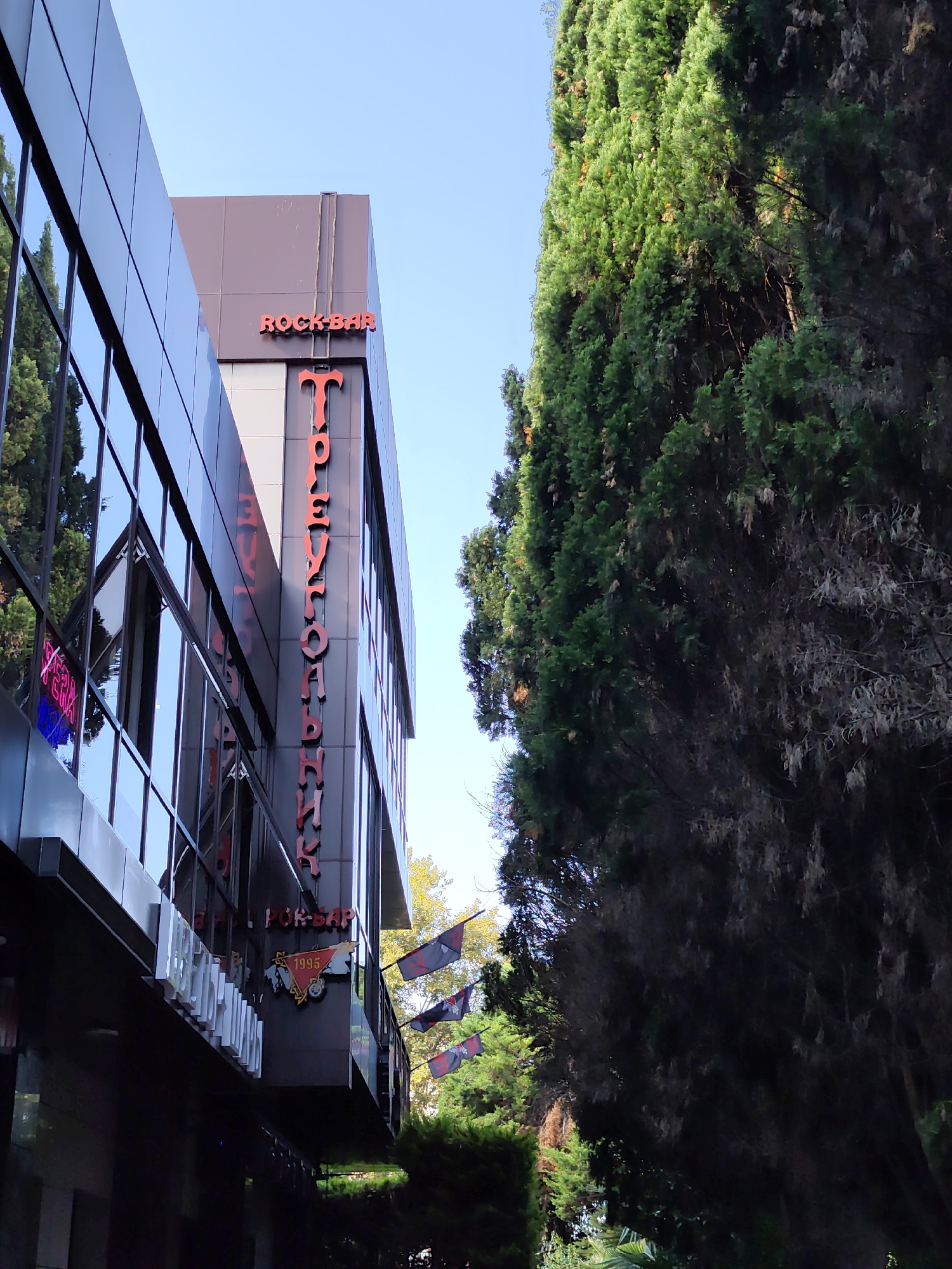 A modern building with a reflective glass facade and dark gray siding is situated on the left side of the image, partially obscured by a large, lush green tree on the right. Red lettering on the building identifies it as "ROCK-BAR". Below this, a larger, vertically oriented red sign displays text that appears to be in Cyrillic, with the letters "Т" and "Р" clearly visible at the top. Further down, a crest-like emblem with the number "1995" and a motorcycle motif is visible. Several dark flags with red symbols are hanging from the side of the building. The sky is a clear blue. The scene is likely located in Sochi, Russia, as suggested by the Cyrillic text and the architectural style.