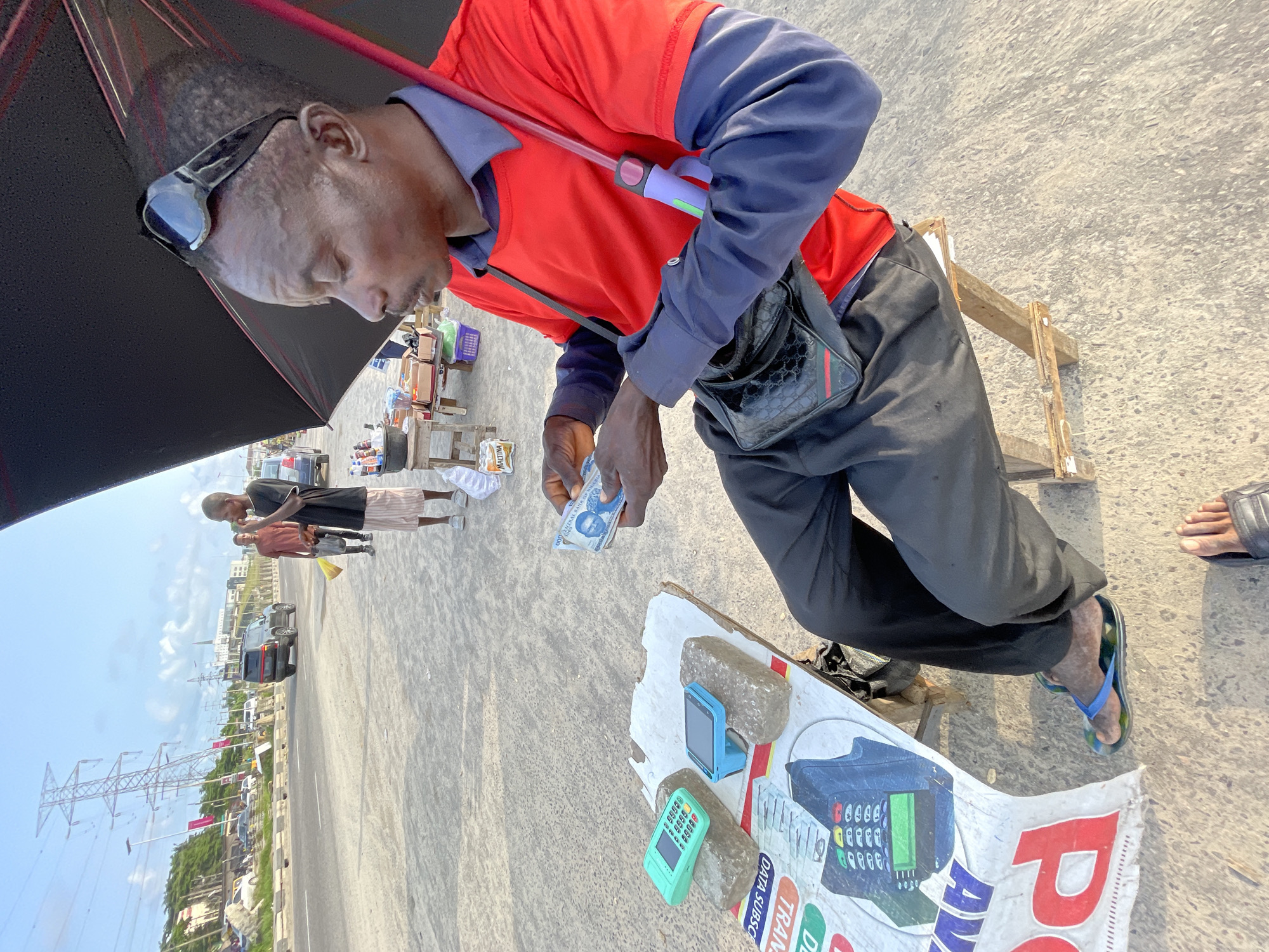 In Lekki, Nigeria, a man sits on a wooden stool under a black umbrella, holding Nigerian currency. He is wearing a red t-shirt over a navy blue collared shirt and black trousers, with blue and green flip-flops. A black patterned crossbody bag is slung over his shoulder. In front of him, on a white surface supported by stones, are two mobile point-of-sale devices, one turquoise and one teal. A large banner with red lettering and an image of a POS machine is visible beneath the white surface. In the background, a paved road is lined with vehicles, including a black SUV, and tall electricity pylons dominate the skyline. Two other men are walking in the distance, one in a maroon t-shirt and striped shorts, and another in a black t-shirt and striped shorts. The ground is dusty and appears to be a roadside market or waiting area.