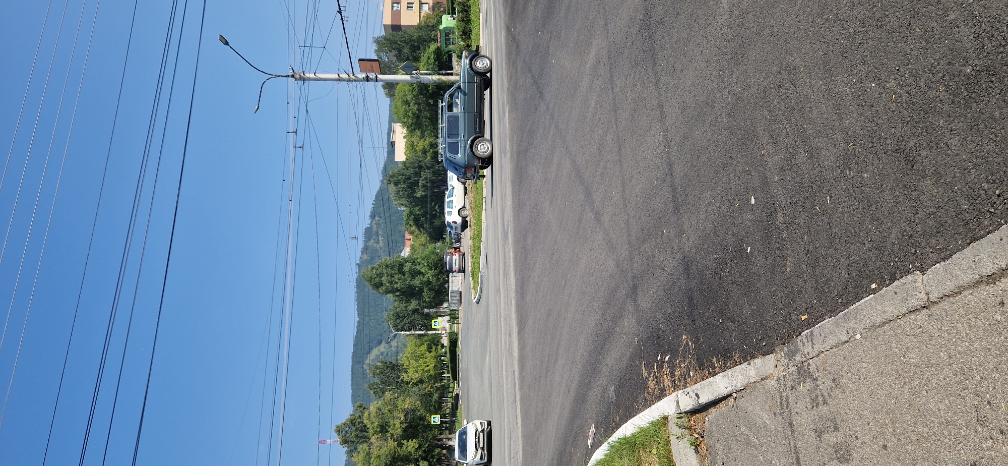 A dark green Lada Niva is driving through an intersection in Miass, Russia. The car has a roof rack. The intersection is paved with asphalt and bordered by a concrete curb with some dry grass and debris. In the background, there are trees, buildings, and a hill. Several other cars are parked or driving in the background. The sky is clear and blue, with several overhead power lines.