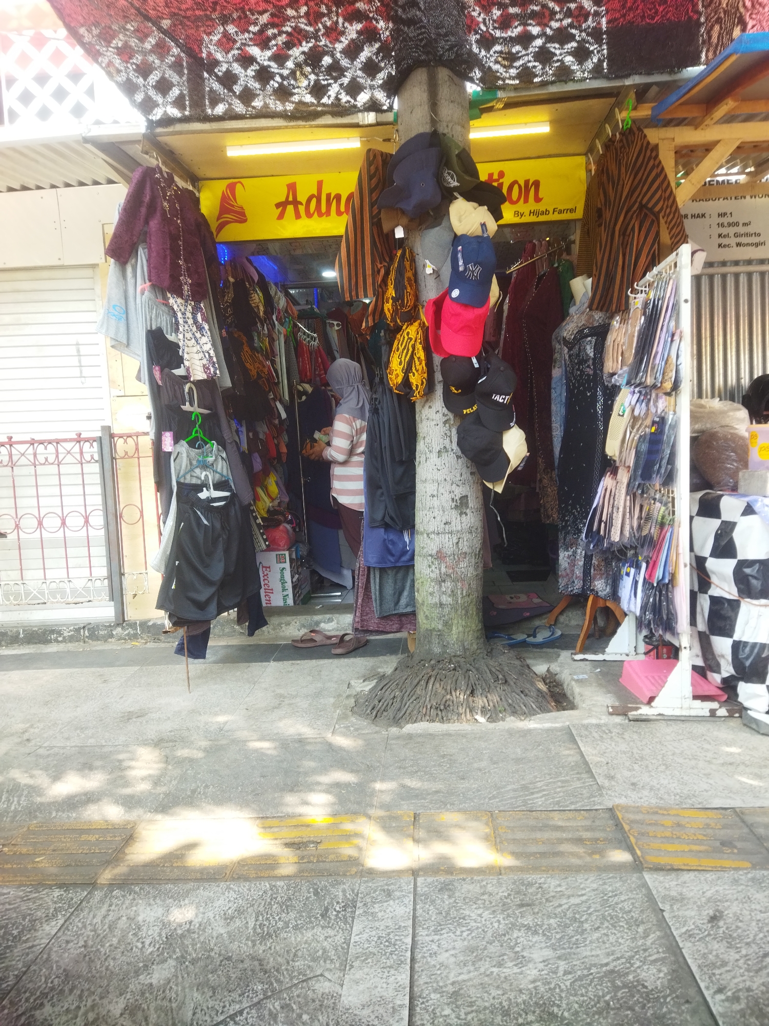 The image depicts an outdoor stall selling clothing and accessories, situated on a sidewalk in Wonogiri, Indonesia. The stall is set up in front of a building with a yellow awning that reads "Adna" and "By: Hijab Farrel." Various clothing items, including shirts, pants, and headscarves, are displayed on racks and hangers. Several hats are also visible, some hanging from a tree trunk that is part of the stall's structure. A woman wearing a hijab and striped shirt is browsing the merchandise. The sidewalk is paved with concrete tiles, and a tactile paving strip, likely for visually impaired pedestrians, is present. In the background, there is a metal gate and part of a building. Sunlight casts shadows on the ground, indicating it is daytime.
