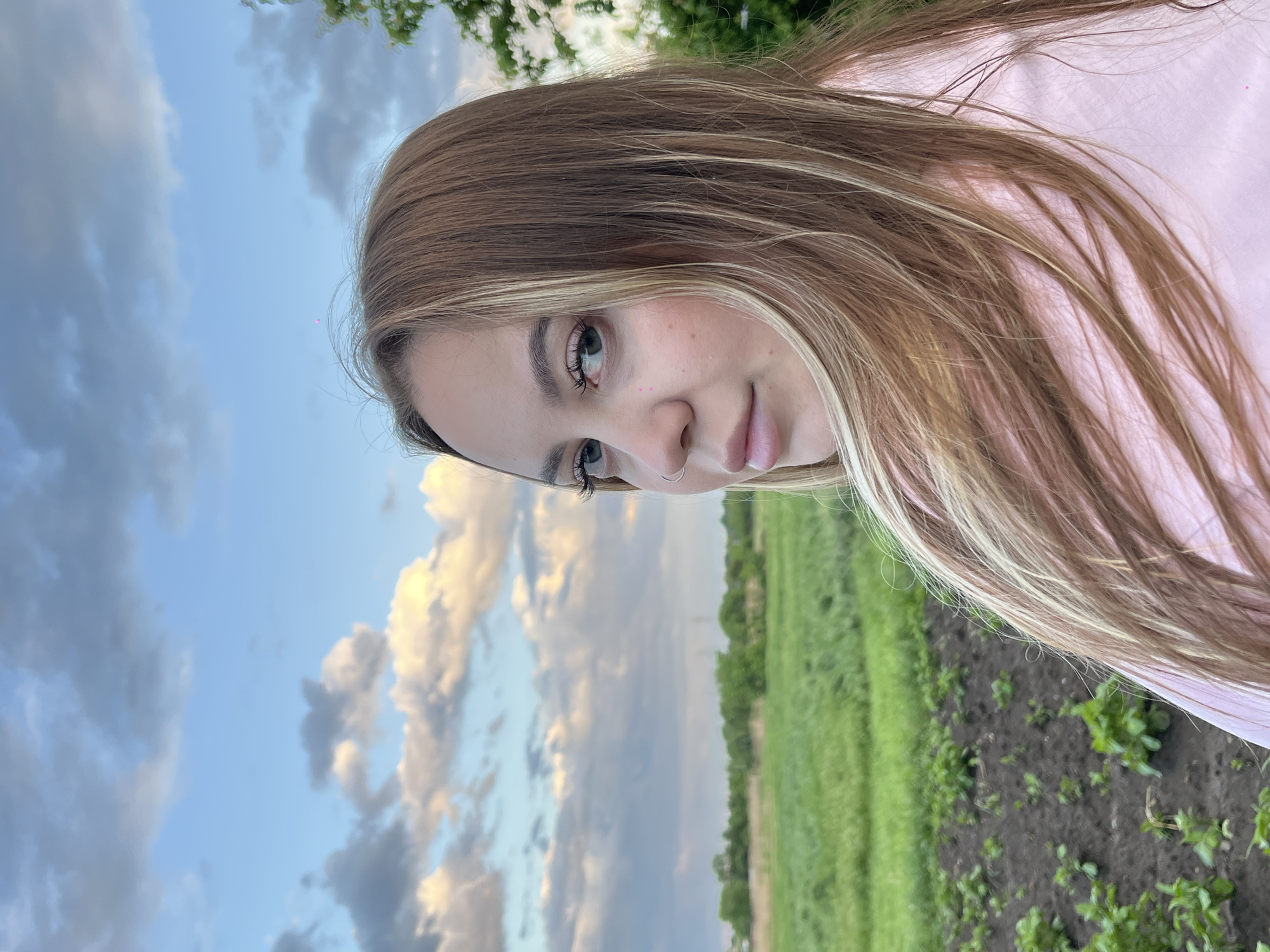 A young woman with blonde hair, wearing a pink top, is photographed outdoors with a field and a cloudy sky in the background. The photograph is taken at an angle, resulting in a landscape orientation with the sky and field appearing vertically. The location is specified as the city of Novopokrovskaya, Russia, but there is no indication of anything city-specific and it mostly seems like a field. The woman is looking slightly to the side with a subtle expression. There are no other people or activities shown in the frame. The only objects visible are the woman, the pink top, the field vegetation, and the sky with clouds.
