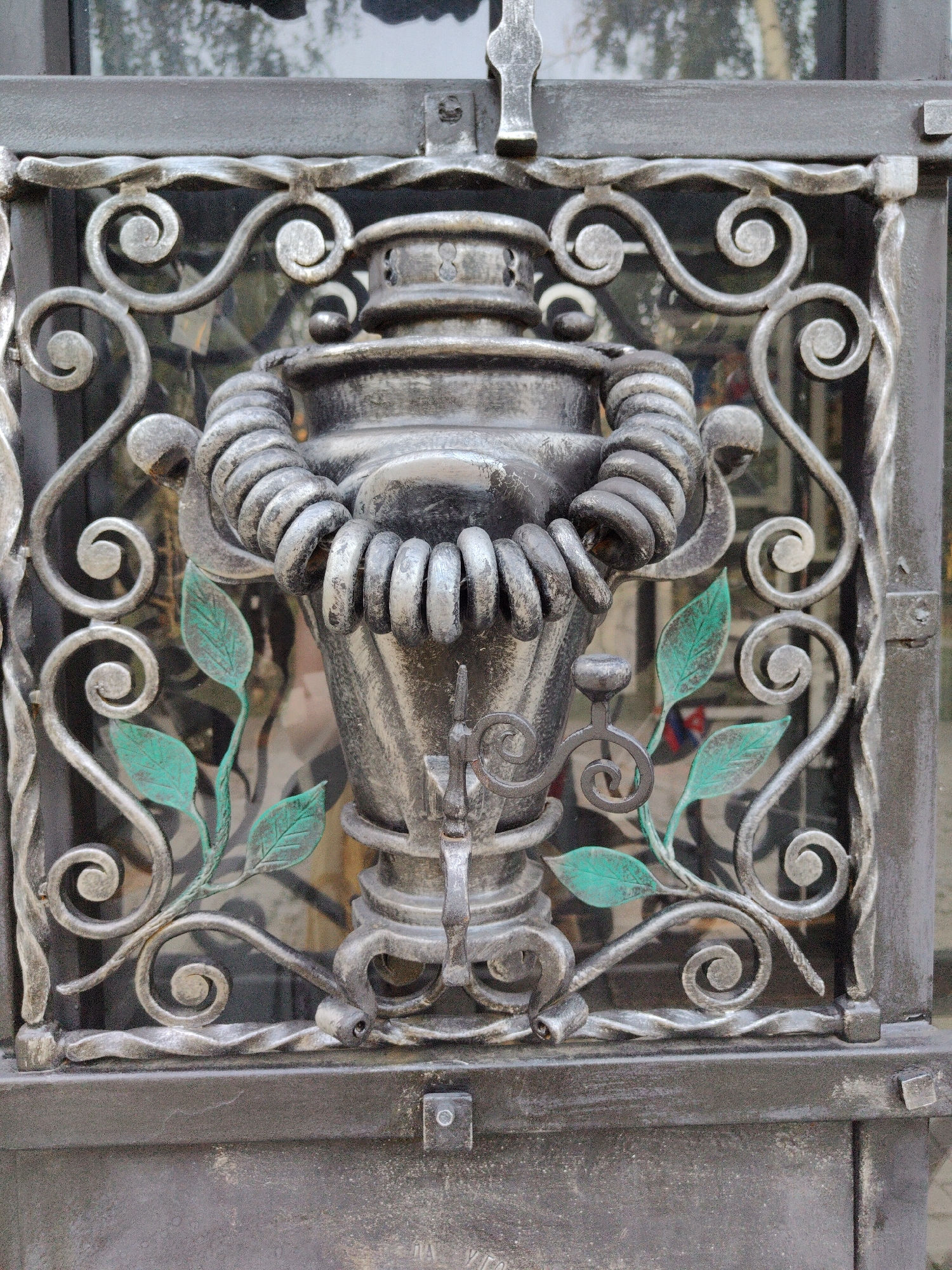 The image shows a close-up, eye-level view of a decorative wrought iron gate in Donetsk, Ukraine. The gate features a central ornate design depicting a samovar, a traditional Russian tea urn, with intertwined metalwork resembling ropes around its handles. Green metal leaves are attached to the scrollwork surrounding the samovar, adding a touch of color to the otherwise gray metal. The background visible through the gate appears to be a reflective surface, possibly glass, showing a blurred, out-of-focus reflection of greenery and possibly buildings. The metalwork is detailed and appears to be aged, with some areas showing texture and wear. The overall impression is one of a finely crafted, possibly historical, decorative element of a gate.