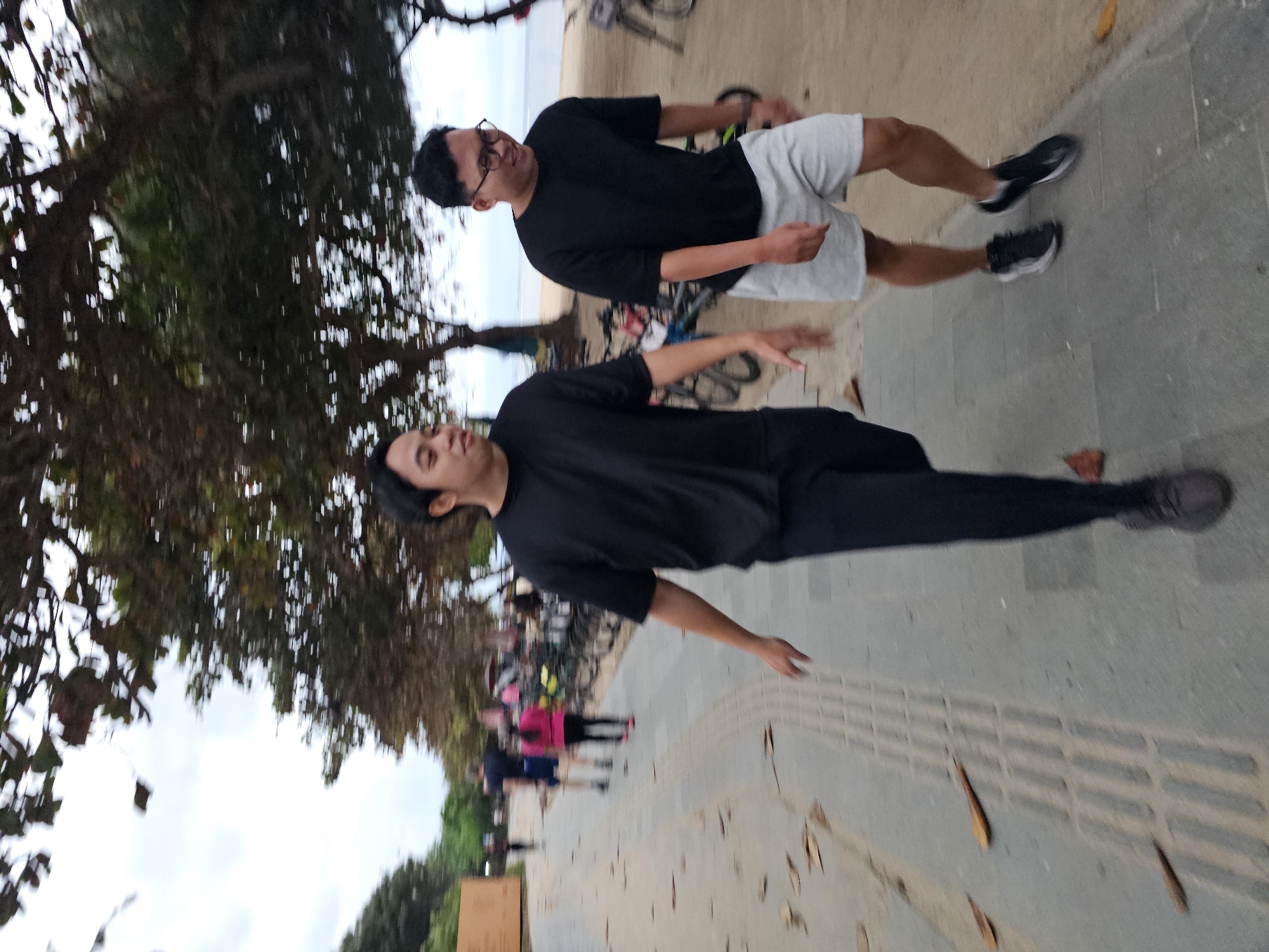 The image depicts a scene along a paved walkway, possibly near a beach in Denpasar, Indonesia. Two young Asian men are the primary subjects, walking from left to right. The man on the left wears a black t-shirt and black pants, with his right arm extended as if in motion. The man on the right is dressed in a black t-shirt and light gray shorts, wearing glasses and a slight smile. Both men are captured mid-stride, indicating movement.
The walkway appears to be made of concrete paving stones with a textured surface, possibly a tactile paving system. Scattered brown leaves are visible on the walkway. To the right of the walkway is a sandy area, suggestive of a beach or coastal path. Beyond the sand, bicycles are parked, and further in the background, trees with dense foliage line the area. The sky is overcast. In the distance, other individuals are seen walking, and a large, light-colored structure is visible to the far left. The overall atmosphere suggests an outdoor recreational area.