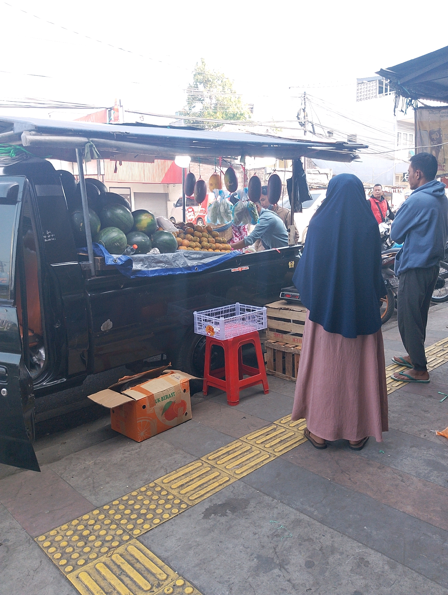 In Lembang, Indonesia, an outdoor fruit stand is set up on a sidewalk. A black pickup truck, with its bed converted into a fruit display, is parked. The bed of the truck is filled with watermelons and oranges. Behind the truck, several people are gathered. A woman wearing a dark blue hijab and a pink skirt stands facing away from the camera, looking towards the fruit stand. To her right, a man in a blue hoodie and dark pants stands with his arms crossed. In the background, a man in a red jacket is visible, appearing to be engaged in conversation or a transaction. A motorcycle is parked near the people. The sidewalk features tactile paving for the visually impaired. A red stool and a white plastic crate are placed on the sidewalk in front of the truck. An open cardboard box with an image of an orange is on the ground next to the truck. The scene appears to be a bustling marketplace.