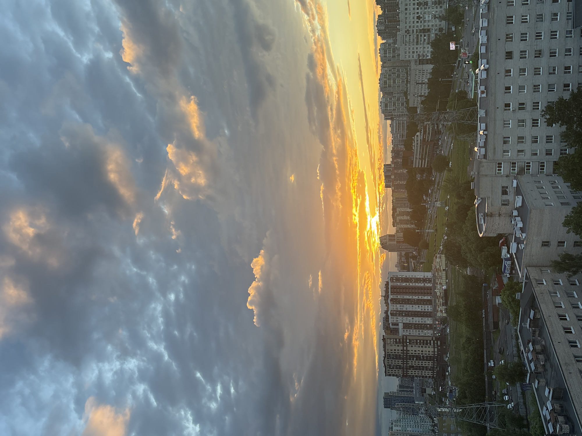 This is a high-angle shot of a city skyline at sunset. The sky is filled with a dramatic mix of dark gray and orange clouds, with the setting sun casting a bright, warm glow. Below the sky, numerous apartment buildings of varying heights and architectural styles dominate the cityscape. Some buildings are modern, with sleek facades, while others appear older and more utilitarian. Green trees and patches of grass are visible between and around the buildings, suggesting parks or unbuilt areas within the urban environment. Power lines and towers are also present, indicating the city's infrastructure. There are no people visible in the frame, and the scene appears to be one of urban development bathed in the atmospheric light of a sunset.