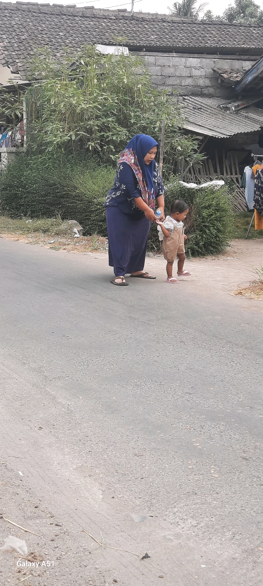 In Klemunan, Indonesia, a woman wearing a blue hijab and a dark blue dress with a floral pattern is standing on an asphalt road. She is holding a clear plastic water bottle and appears to be offering it to a small child. The child, who is wearing tan overalls and pink sandals, is standing facing the woman. Behind them, there is dense green foliage and a building with a tiled roof and gray concrete walls. Clothes are hanging on a rack to the right of the building. The road shows signs of wear and tear, with patches of lighter and darker asphalt. The overall scene depicts an everyday moment in a rural or semi-rural Indonesian setting.