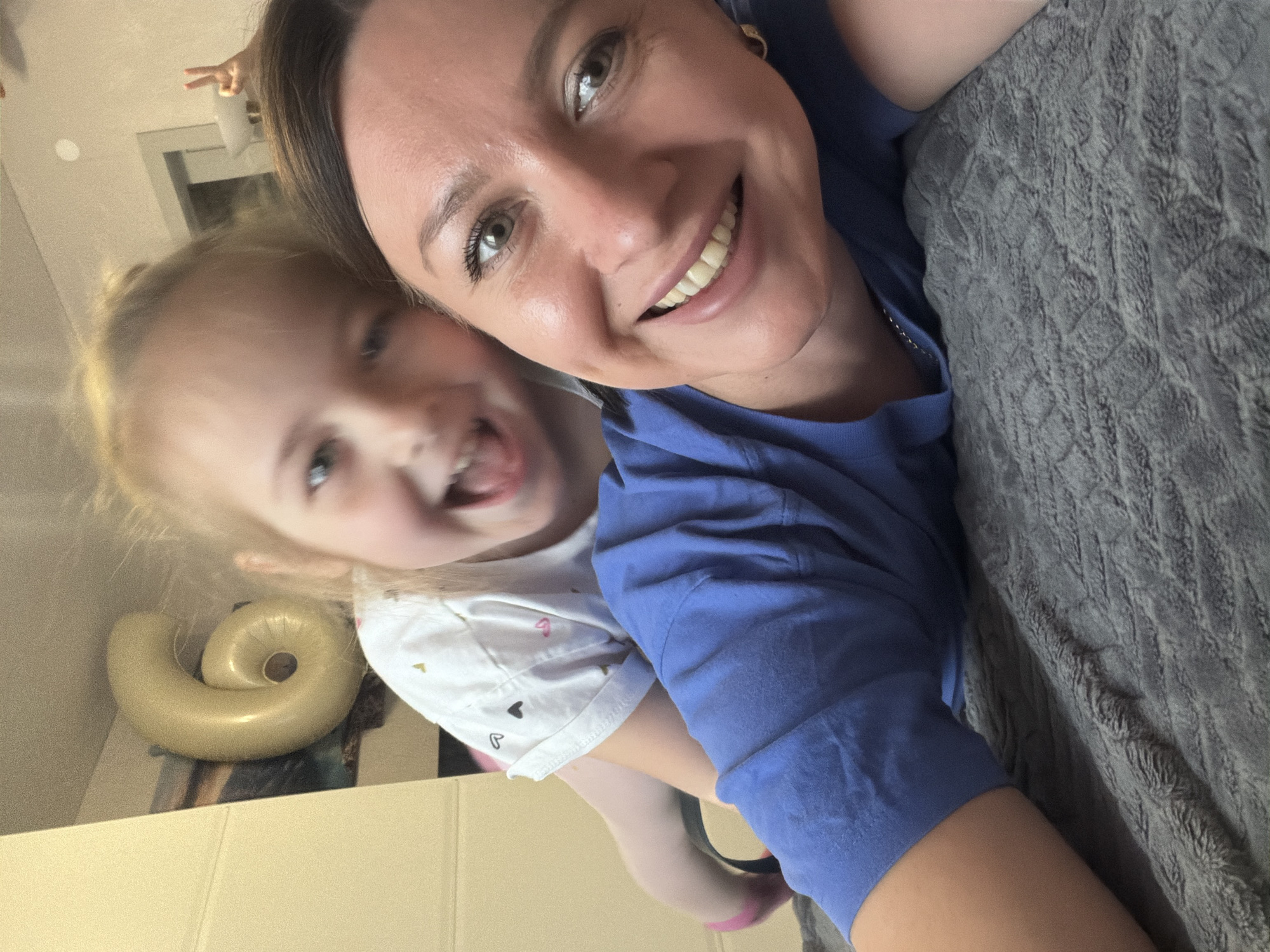 A selfie captures a fair-skinned adult woman and a fair-skinned child. The woman is smiling, her face close to the camera, wearing a blue short-sleeved shirt and what appears to be small, gold-colored earrings. The child is wearing a white shirt with heart patterns, sticking their tongue out and showing a smile. A gray blanket or soft surface fills the right side of the frame. In the background, a neutral-colored wall, a section of a window, and an inflatable pool toy shaped like a snail can be seen. Based on the content, location information is not possible.