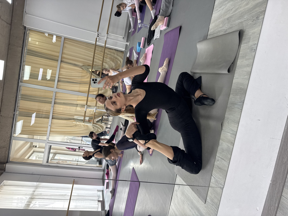 An indoor dance studio in Unknown, Unknown, shows approximately 10-12 females, ranging from young children to adults, engaged in stretching and ballet exercises. In the foreground, an adult female in a black long-sleeved leotard, black pants, and black ballet flats performs a modified King Pigeon yoga pose on a grey mat, holding her left foot with her left hand while taking a selfie with a white smartphone in her right hand. Her reflection is visible in the large mirror covering the left wall of the studio. Other participants are distributed across the light grey floor, performing various stretches on purple yoga mats, some utilizing small blue and pink yoga blocks for support. Attire includes leotards (black, white), tights (pink, white), and ballet flats (pink, black). A wooden ballet barre runs along the far wall, where large windows with light beige curtains provide natural illumination. White radiators are mounted beneath the windows. The studio features high ceilings, partly exposed concrete, and a speaker on the right wall.