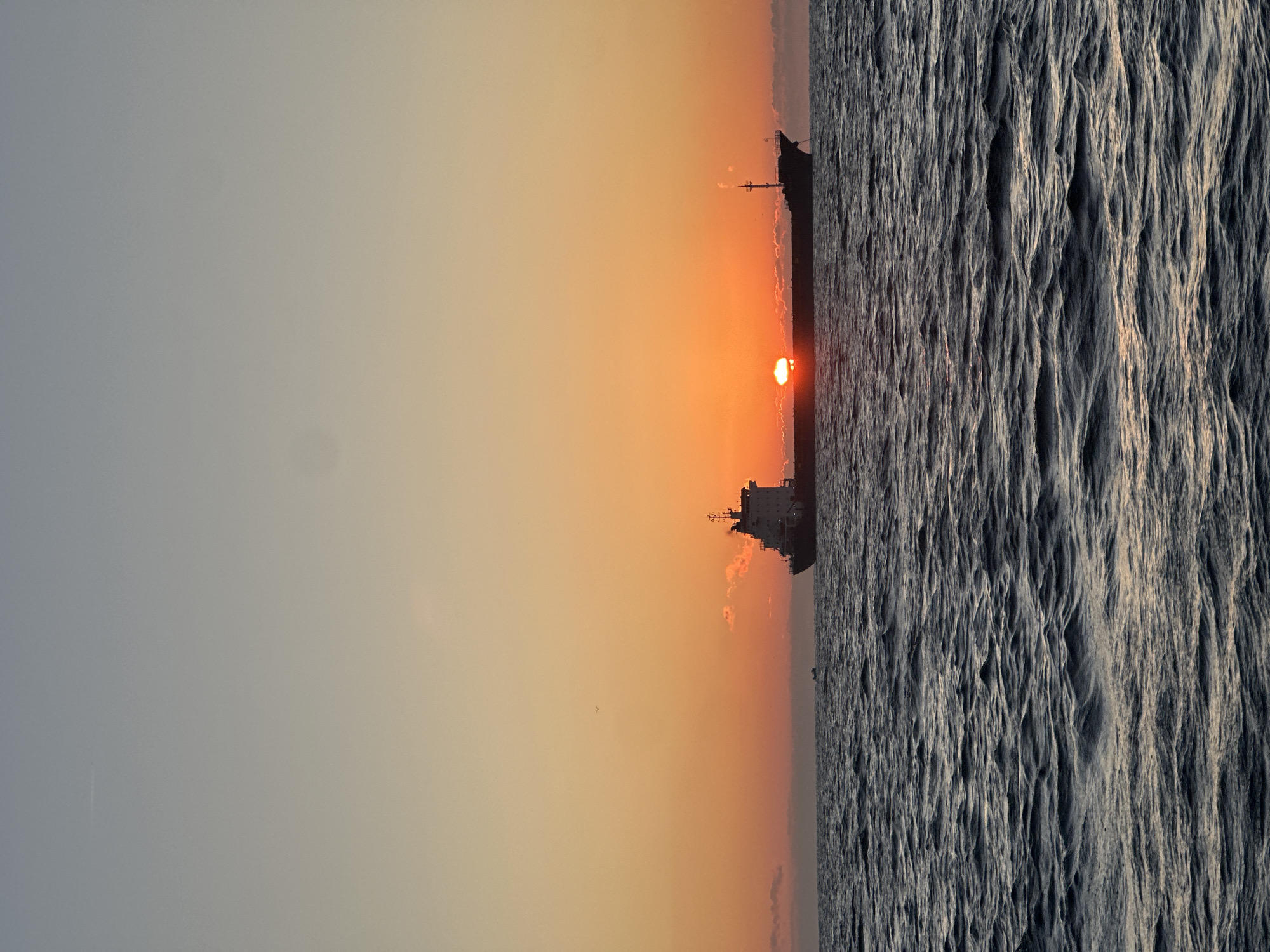 An immense cargo ship is silhouetted against a vibrant sunset over a dark, choppy ocean. The sun, a brilliant orange orb, is partially obscured by a thin layer of clouds as it dips towards the horizon. The sky transitions from a pale, hazy blue at the top to a warm orange, then a deep red near the water. The ocean surface is a deep, dark blue, with white crests on the waves reflecting the fading light. The ship, a long, dark shape, dominates the middle ground, with its superstructure clearly visible. In the distance, a much smaller vessel is also visible on the water. The scene is tranquil and expansive, evoking a sense of both scale and serenity.
