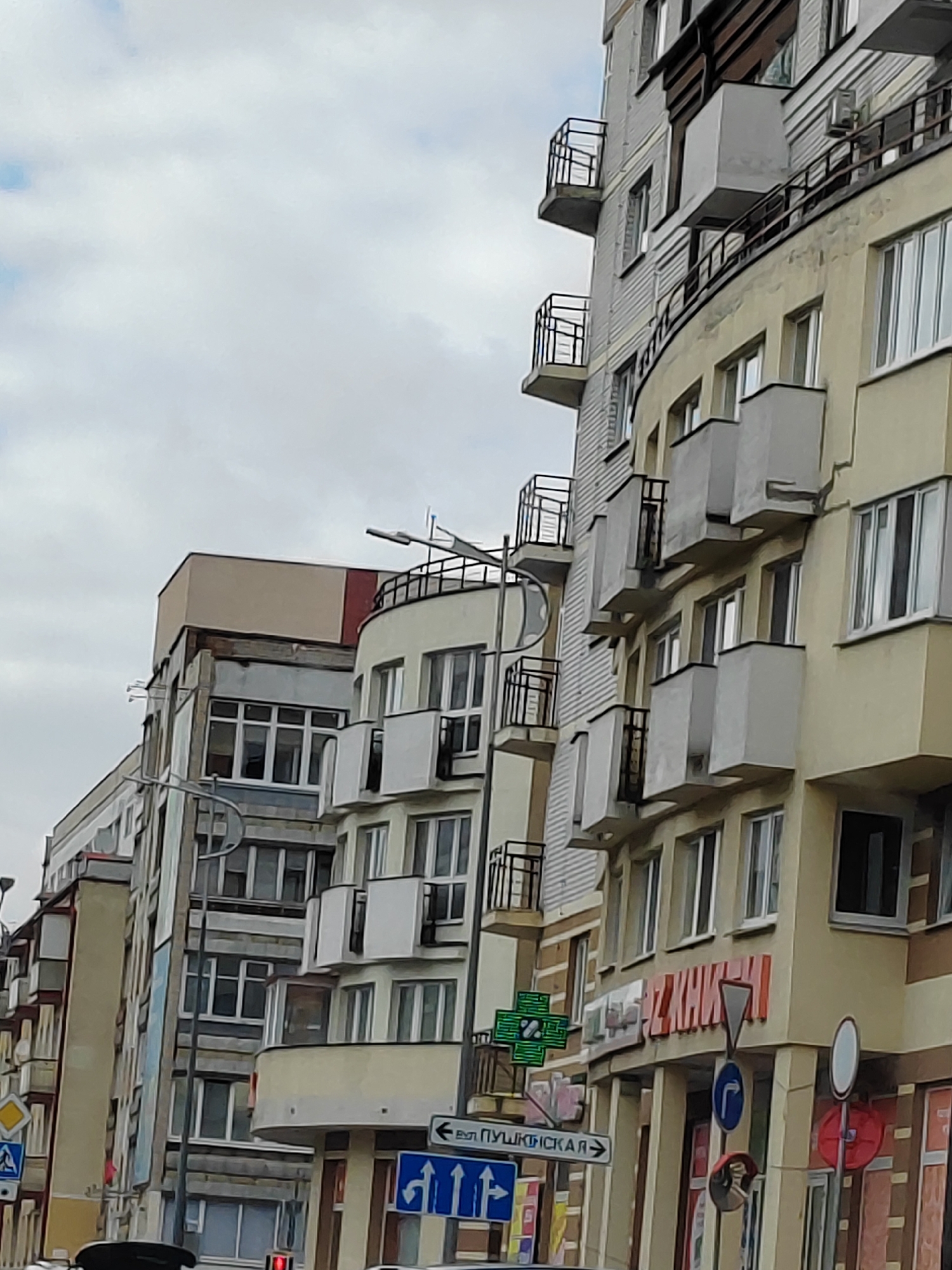 The image shows a street view in the city of Brest, Belarus, featuring a row of multi-story buildings with various architectural styles. The buildings are predominantly light-colored, with yellow and beige facades, and some exhibit curved designs. Several balconies with metal railings are visible. Street signs and a traffic light can be seen, indicating an urban setting. A green cross symbol, commonly associated with pharmacies, is displayed on one of the buildings. The sky is overcast with clouds. The overall scene depicts a typical street in a European city.