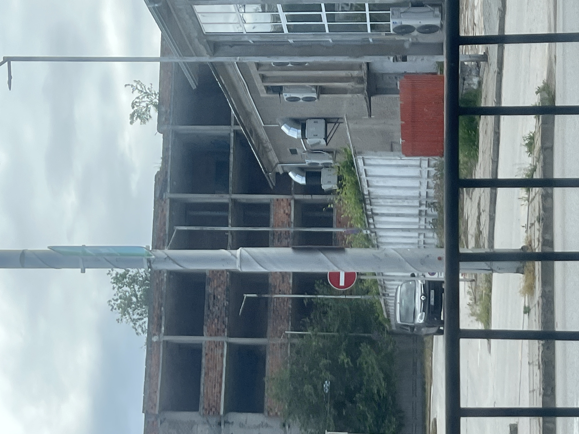 The image shows an exterior view of a partially constructed or abandoned building in Plovdiv, Bulgaria. The building has multiple floors with open, unglazed window frames revealing brickwork. Air conditioning units are affixed to the building's exterior. There is vegetation growing near the building. Street signs and a car are visible through metal bars. A "Do Not Enter" sign is partially obscured by foliage. The sky is overcast.