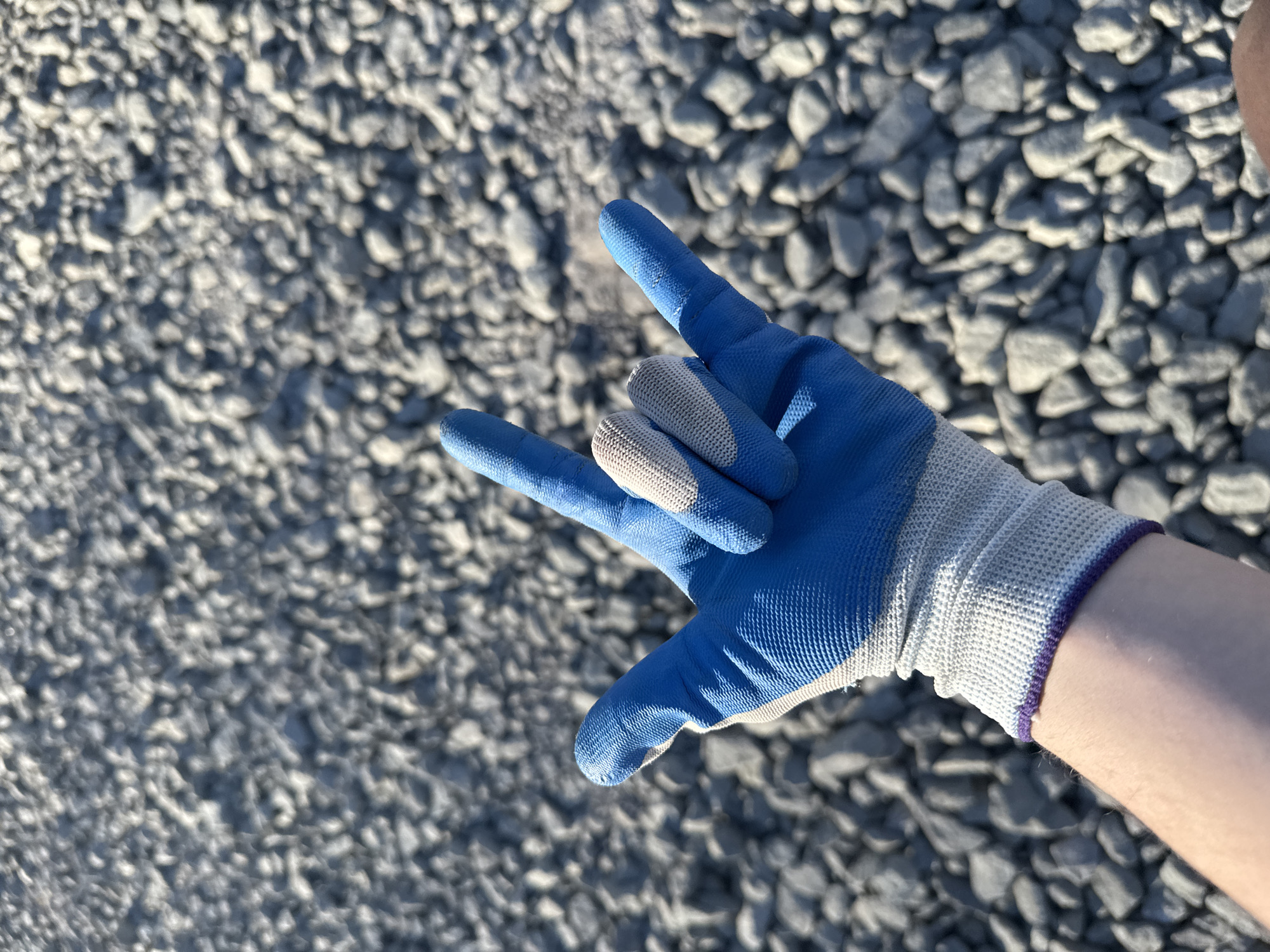 A gloved hand, making the "I love you" sign, is positioned against a background of gray rocks. The glove is blue with gray accents on the fingers and wrist. The arm of a person wearing the glove is visible. The setting is a gravelly, rocky ground surface. There is no specific indication of the location being Kazan, Russia.