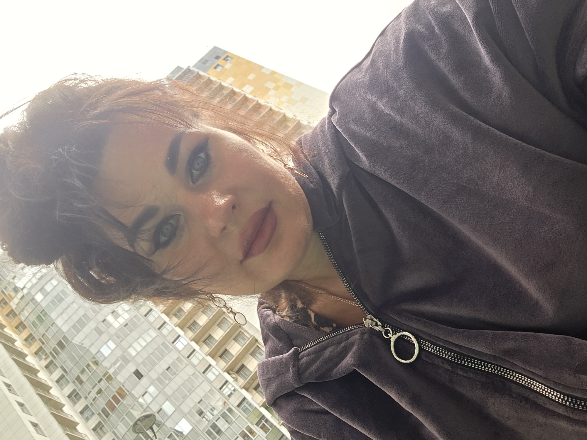 A woman with her hair in a bun and wearing a dark grey zip-up hoodie stands in front of a modern building in Saint Petersburg, Russia. The building appears to be a residential or office complex with numerous windows. The woman is looking at the camera and has makeup on. She is also wearing earrings and a necklace. The image is taken from a low angle, looking up at the woman and the building. The lighting suggests it is daytime.