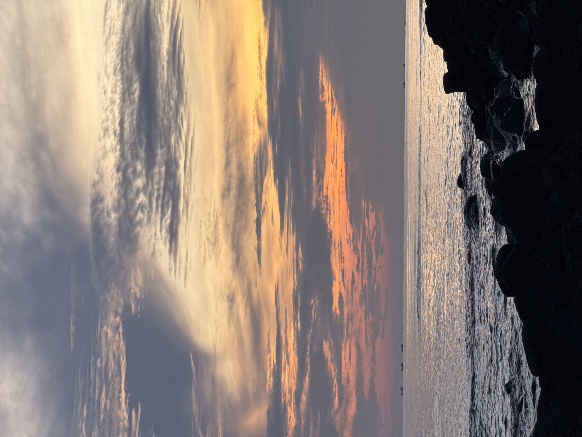 A serene seascape is captured at dusk on Lý Sơn Island in Vietnam. The sky is a canvas of blended colors, transitioning from a soft yellow at the horizon to muted oranges and pinks, with streaks of grey clouds creating a textured effect. Below the sky, the calm sea reflects the fading light, with gentle waves lapping against a rocky shore in the foreground. The rocks are silhouetted against the water, their dark forms contrasting with the softer hues of the sea and sky. In the distance, three small boats are visible on the horizon, adding a sense of scale and human presence to the vast natural expanse.