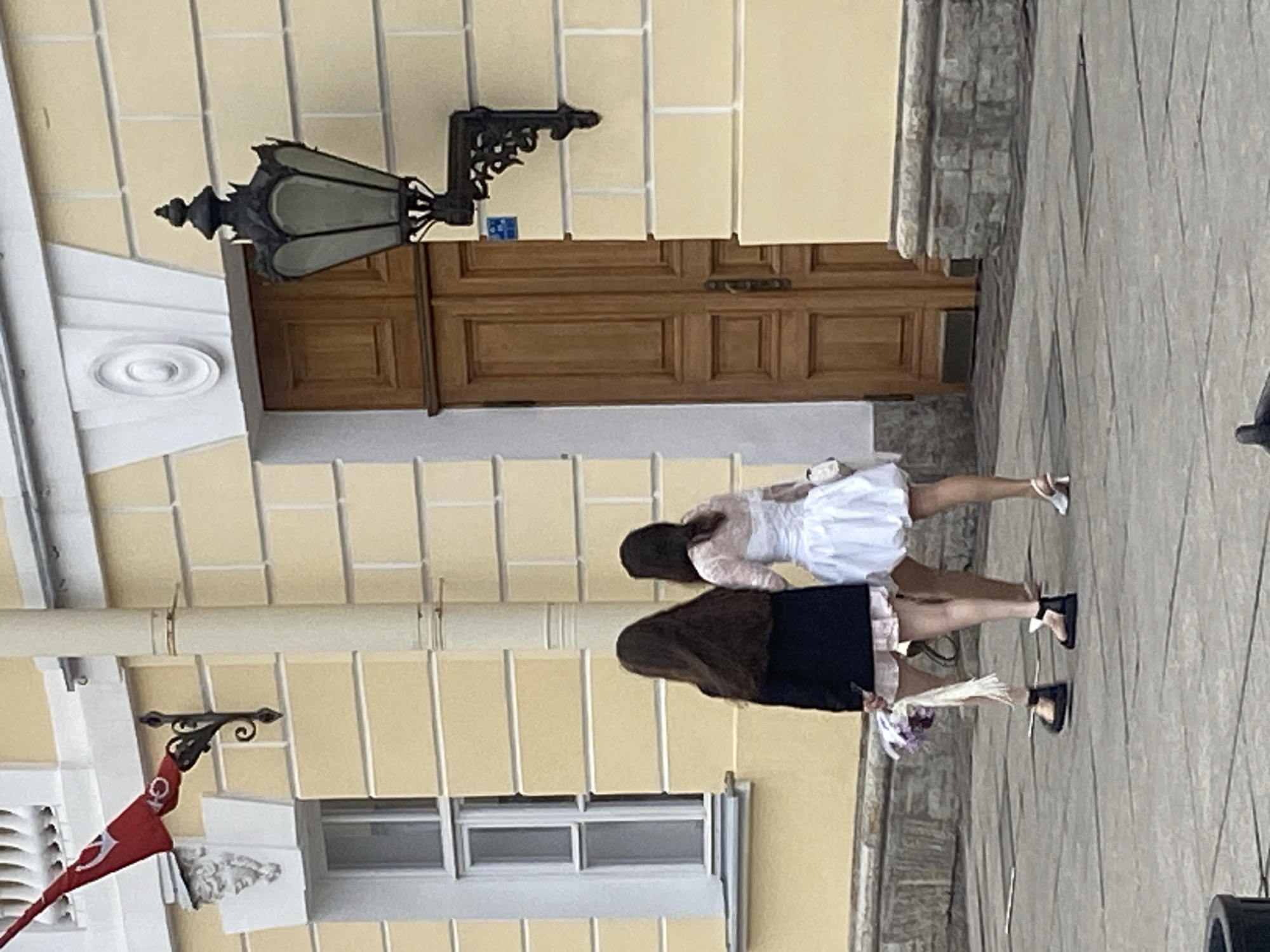 Two young women, viewed from behind, walk across a stone-paved area in front of a light yellow building with a large wooden door and a tall window. The woman on the left wears a dark jacket over a light-colored skirt and dark sandals. She carries a bouquet of flowers. The woman on the right wears a white, short dress with puffed sleeves and white heeled sandals. She carries a small white purse. A red flag with a white symbol is visible mounted on the building to the left, near a decorative architectural element. A vintage-style black lantern is mounted on the building to the right of the door. The building's facade features decorative white molding above the window and door. The scene appears to be in Saint Petersburg, Russia, given the architectural style and the presence of a flag.