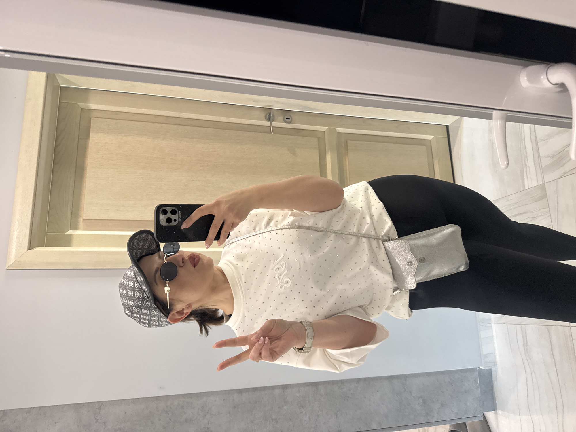 A woman is taking a selfie in a bathroom. She is wearing a patterned cap, sunglasses, a white shirt with small embellishments, black leggings, and a silver cross-body bag. She is holding up a peace sign with her left hand. The bathroom has a light wood door, white walls with gray accents, and light marble-patterned floor tiles. The location is in the city of Sofia, Bulgaria, though specific information of the surrounding environment is not available.