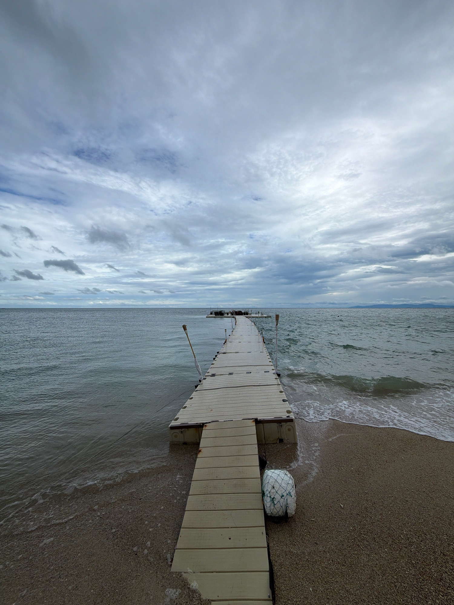 A long, light-colored wooden pier extends into a calm, grey body of water under a cloudy sky. The pier is constructed of modular sections, creating a slightly uneven surface. Small waves lap at the sandy shore where the pier begins. A white, mesh-covered ball sits on the sand near the base of the pier. A single tiki torch on a pole is attached to the side of the pier. In the distance, the water meets a hazy horizon, and a faint outline of land is visible. The overall scene is subdued and overcast. The location is not identifiable as Ko Samui District, Thailand based on the visual information provided.
