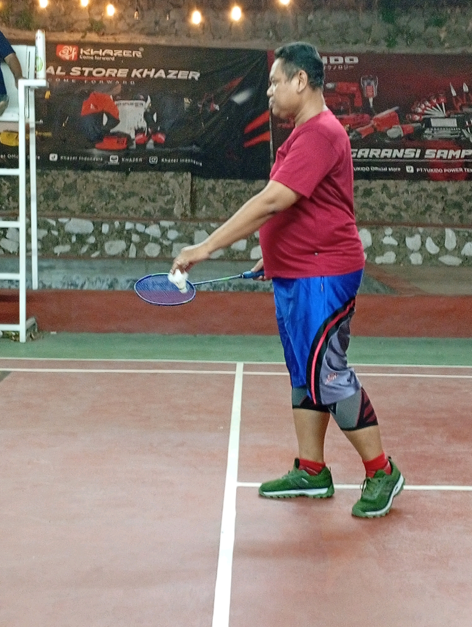 A full-body, side-profile shot shows a medium-built man standing on a badminton court in Caringin, Indonesia. The man is wearing a maroon t-shirt, blue shorts with a black and red design, knee-high grey and red compression sleeves, red socks, and green athletic shoes. He is holding a badminton racket in his right hand, with a white shuttlecock positioned above it, indicating he is about to serve or has just served. The court is made of a reddish-brown material with white boundary lines. In the background, a large banner with text and images is visible, along with a stone wall and some outdoor lighting.
