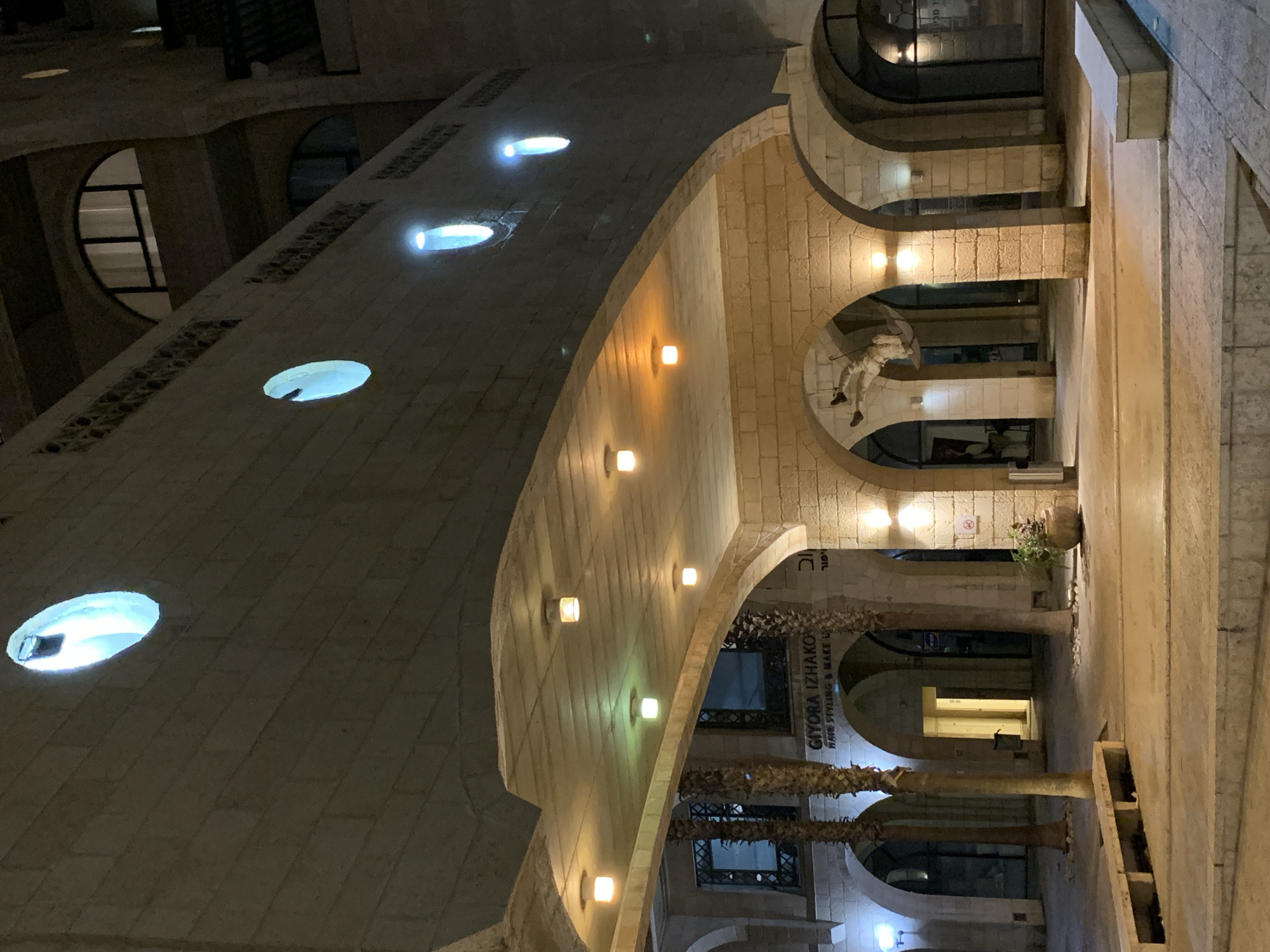 This nighttime photograph depicts a dynamic architectural scene, likely a pedestrian walkway or commercial area, characterized by stone structures and artificial lighting. The image is captured from a tilted perspective, emphasizing the diagonal lines of the building elements.
In the upper left, a long, angled canopy or ceiling, constructed from light-colored stone blocks, features multiple bright, circular recessed lights emitting a cool blue-white glow. Below this, a curved stone wall is illuminated by a series of square, wall-mounted fixtures that cast a warm, orange-yellow light.
The architecture transitions into a series of classic stone arches, supported by columns, in the middle and right sections of the frame. These arched passages are well-lit by numerous warm-toned lamps. Within one of the arches on the right, a notable art installation is visible: a white, human-like figure hangs, holding an open umbrella.
Several tall palm trees with rough trunks extend upwards from the lower right, appearing to line the arched pathway. Behind the arches, dark windows or glass storefronts reflect the ambient light, and a small potted plant can be seen on a ledge.
Visible text on a sign within one of the arched sections clearly reads "GIYORA ITZHAKOV" in English, with "HAIR STYLING & MAKE UP" beneath it, indicating a salon or similar business. Corresponding Hebrew text "גיורא יצחקוב" is also present. Other, less legible text appears etched into the upper stone structure.
The scene is devoid of people engaged in activity, focusing instead on the illuminated architecture and art installation. The extensive use of artificial lighting confirms the time of day as night, creating a dramatic and inviting atmosphere. The architectural style, palm trees, and visible Hebrew text align with the provided location context of Tel-Aviv, Israel.