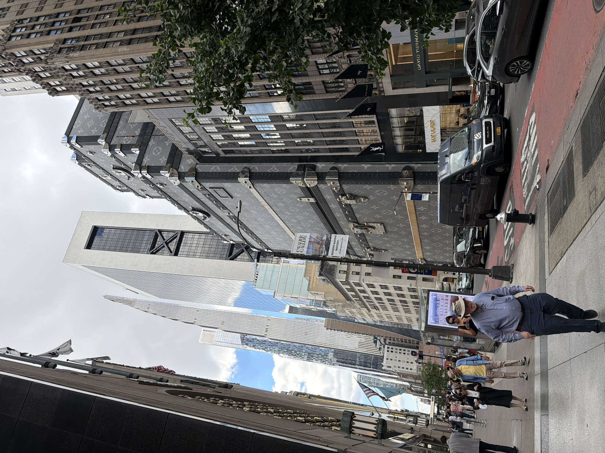 This image depicts a street view in New York City, specifically on Fifth Avenue, between 57th and 58th Streets, a well-known luxury shopping district. The perspective is from the sidewalk looking down the street, with tall buildings on both sides.
In the foreground, a man in a blue checkered shirt and dark pants, wearing a straw hat, walks towards the camera on the sidewalk. Several other pedestrians are visible on the sidewalk behind him, walking in both directions. Some are dressed in casual attire, while others appear more formally dressed.
On the right side of the street, a large, dark-colored van is parked in traffic. Several cars, including a dark sedan and a silver hatchback, are also in motion or stopped in the street. The street is marked with red lanes.
The buildings lining the street are prominent. On the left, a section of a building with dark stone and gold accents is visible, along with an ornate street lamp. On the right, a building is covered in a giant Louis Vuitton trunk motif, featuring the brand's signature monogram and large metal buckles. Adjacent to this, the storefronts of designer boutiques like Saint Laurent and David Yurman are visible. Further down the street, several modern skyscrapers with glass facades dominate the skyline. The sky is cloudy with patches of blue.
An American flag and other flags are displayed on poles outside some of the buildings. Promotional banners and signs are also visible on the street and buildings, including one for "Cavalier Gallery" advertising "Modern Marine Masters." The scene suggests a bustling urban environment with a mix of pedestrian and vehicular traffic, set against a backdrop of impressive architecture and high-end retail.