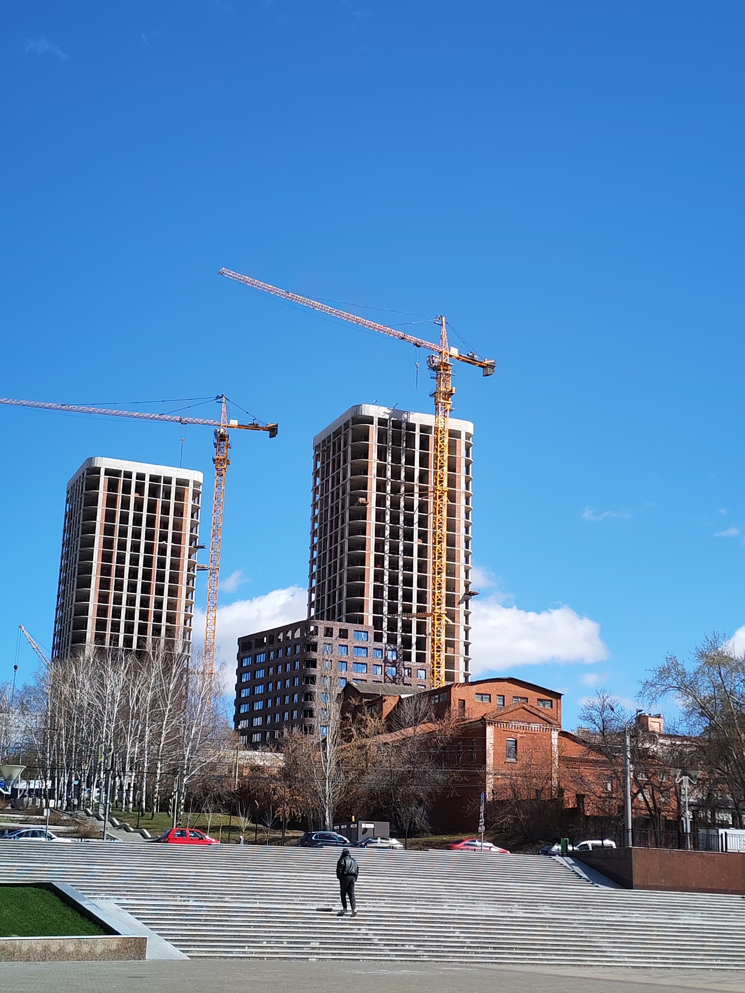 The image shows an urban scene in Izhevsk, Russia, dominated by two tall, unfinished concrete buildings under construction. Each building is accompanied by a yellow crane. In front of the buildings is a multi-level staircase. A person in a black jacket is standing on the stairs. Behind the buildings are other structures, including a red brick building with a distinctive roofline. Bare trees are interspersed in the scene. Several cars are visible in the background. The sky is clear blue with scattered clouds.