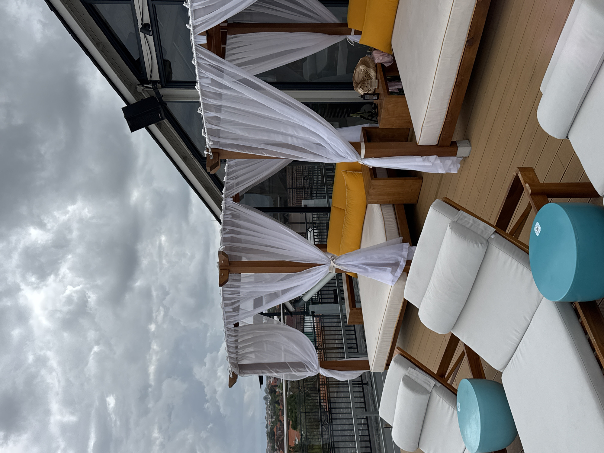 The image depicts a rooftop lounge area in North Kuta, Indonesia, featuring two white canopied daybeds with yellow cushions. In the foreground, two white lounge chairs with blue cylindrical side tables are visible, positioned on a wooden deck. The background reveals a cityscape with buildings and a cloudy sky. A speaker is mounted on the sloped roof of an adjacent building.