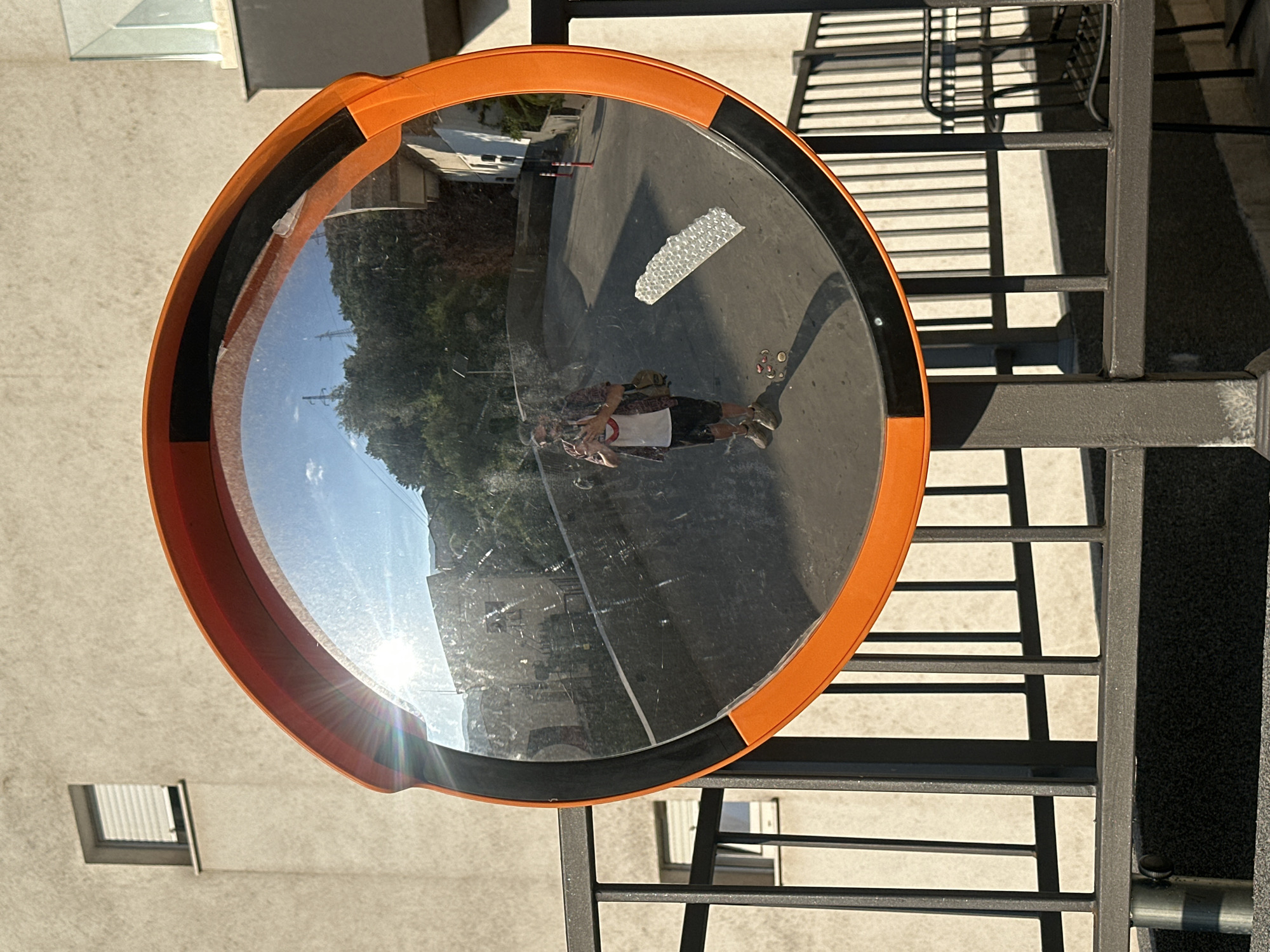 The image is a reflection in a convex mirror, positioned outdoors, likely in the city of Budva, Montenegro, given the architectural style and lush greenery. The mirror itself has an orange frame with black trim. It is mounted on a dark gray metal post, which is attached to a dark gray railing. Behind the mirror, a light-colored building with a small, square window is visible. Further in the background, trees and a road can be seen.
The reflection in the mirror shows a person taking the photograph. This person is wearing a patterned short-sleeved shirt, a white t-shirt underneath, dark shorts, and light-colored shoes. They are holding a smartphone in their hands, with the camera facing them. A light-colored messenger bag is slung across their body. In front of the person in the reflection, a small, light-colored object with wheels, possibly a toy or a skateboard, is on the ground. A piece of clear plastic, appearing to be bubble wrap, is affixed to the road surface in the reflection. The sun is shining brightly, creating lens flare on the left side of the mirror and casting shadows on the ground. The sky is blue with some clouds. The overall scene suggests a sunny day in an urban or semi-urban environment.